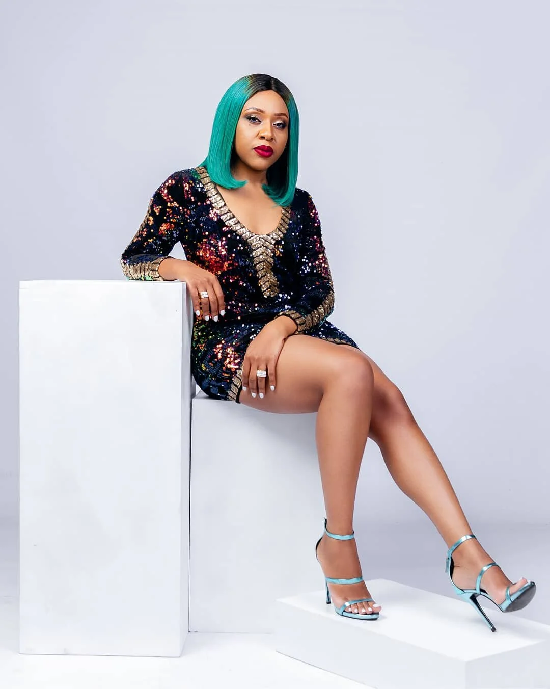 A woman with shoulder-length teal hair, wearing a multi-colored sequined dress and teal high-heeled sandals, sitting on a white platform against a plain gray background.