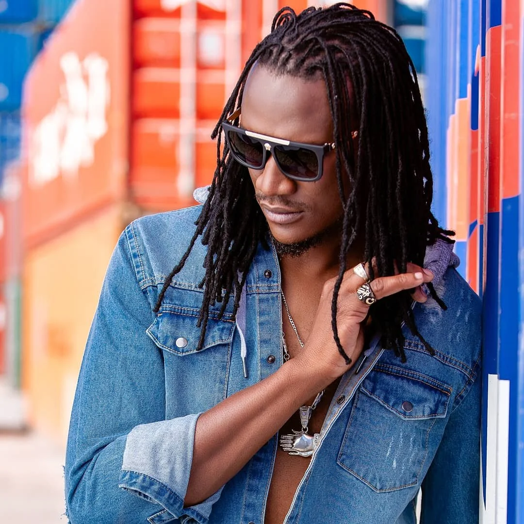 A man with long dreadlocks wearing sunglasses and a denim jacket, standing against colorful lockers.