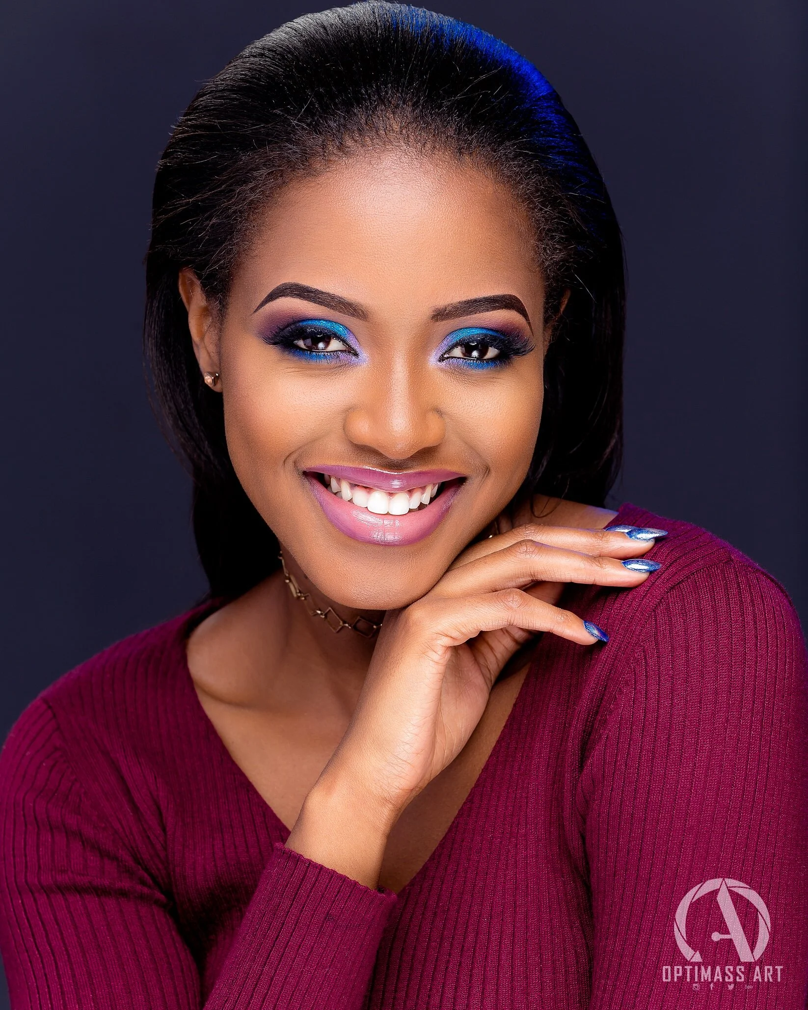 A smiling woman with dark hair, vibrant blue eyeshadow, pink lipstick, wearing a maroon top and a choker necklace, posing against a dark background.