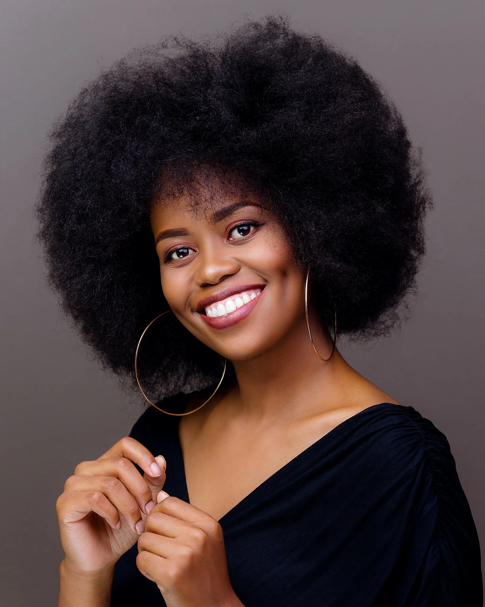 Portrait of a smiling African American woman with big curly hair, wearing hoop earrings and a black top against a gray background.