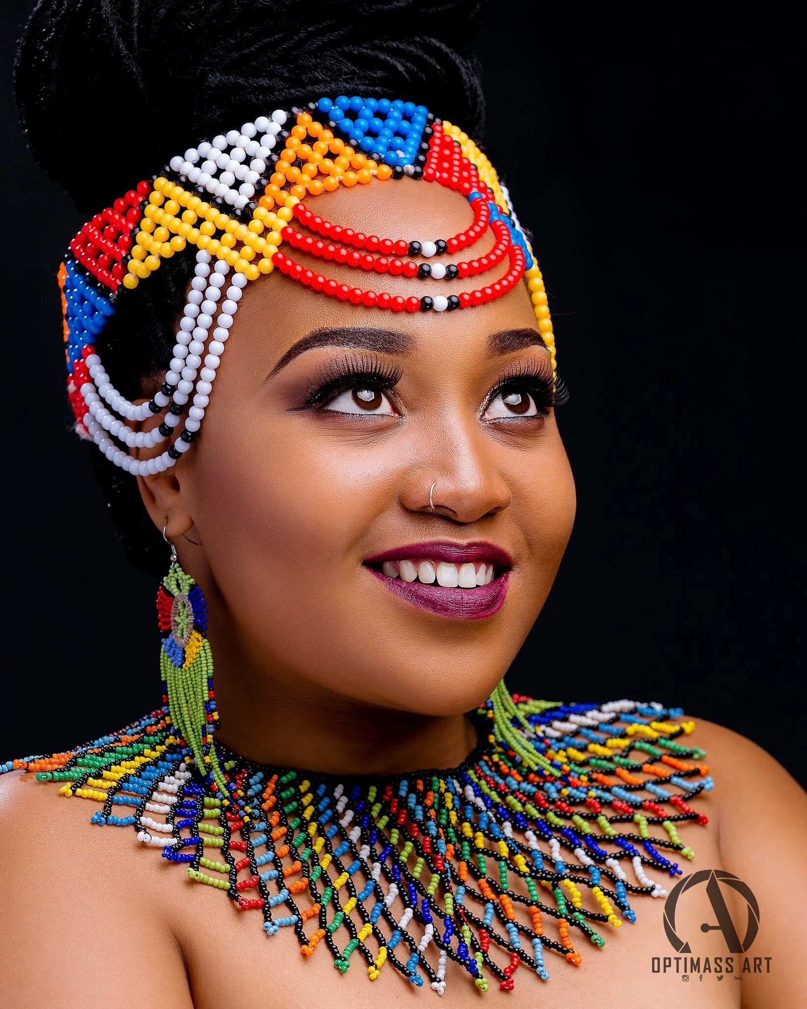 A woman with a colorful beaded headwrap, earrings, and necklace, smiling and looking up, against a black background.