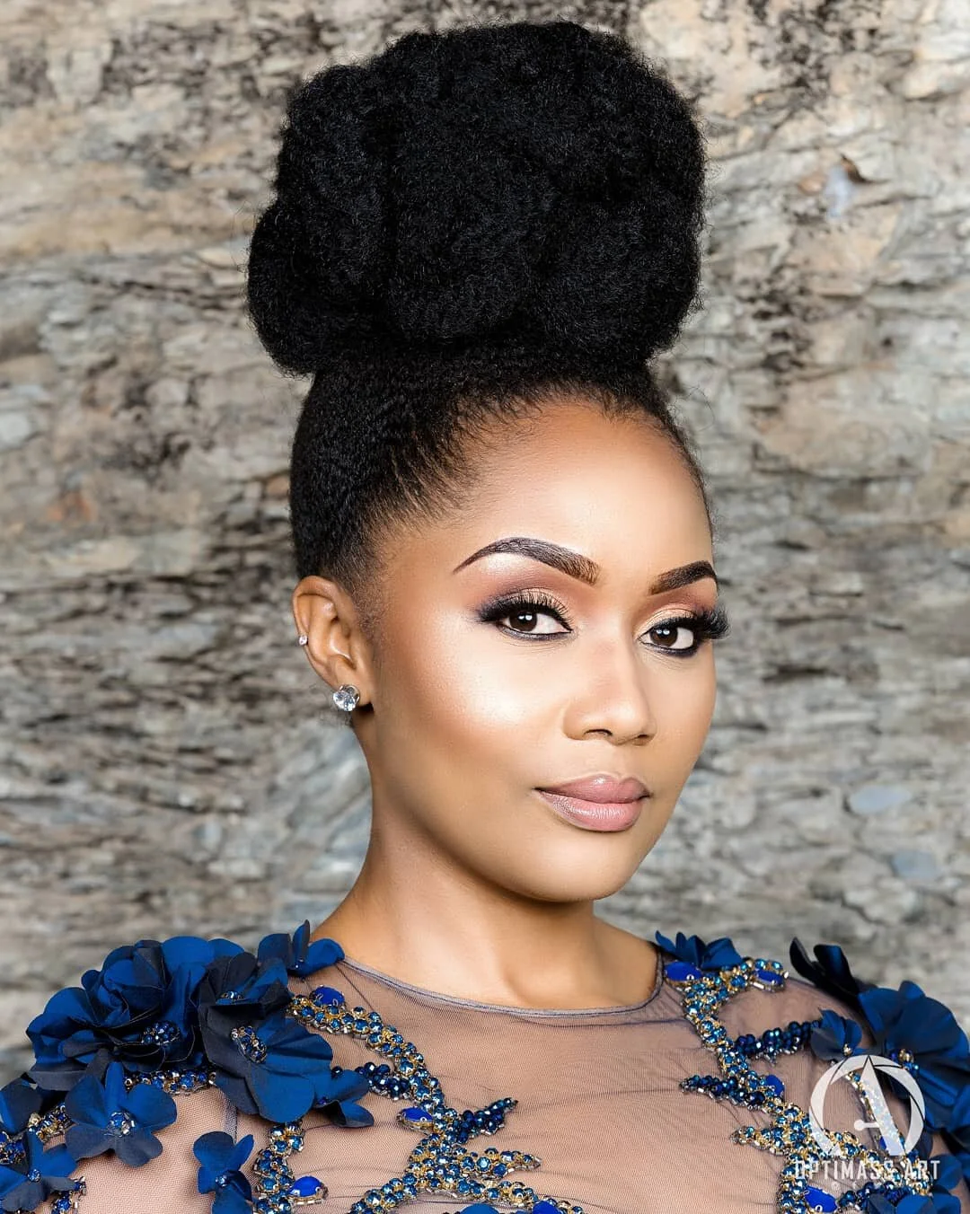 A woman with a high, large braided bun hairstyle, wearing makeup and diamond earrings, dressed in a blue and beige dress with floral embellishments, standing against a stone wall background.