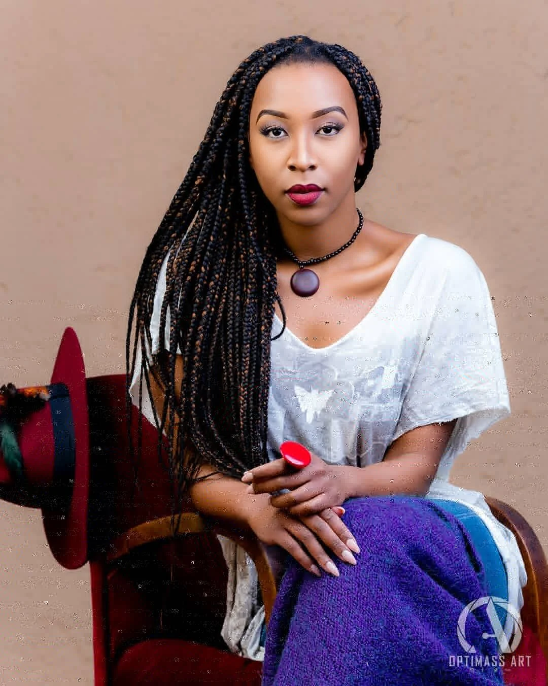 A woman with long braided hair, wearing a white top, black choker, and a large purple pendant, sitting with her hands folded and a red and purple object, possibly a cushion or blanket, in her lap, against a beige background.