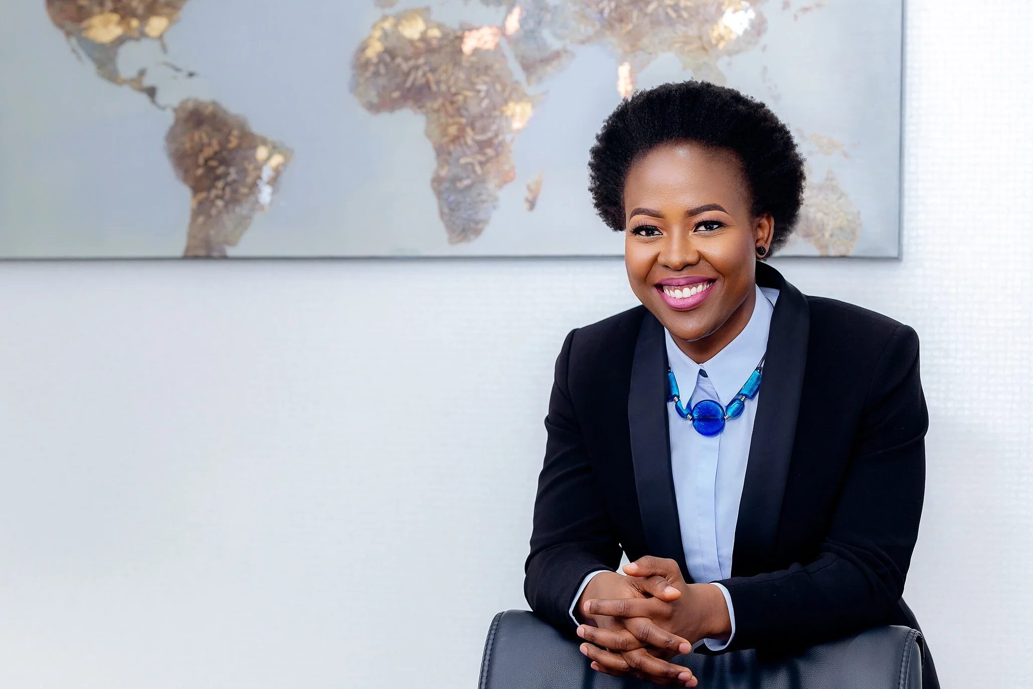 A smiling woman in a business suit with a blue and black necklace, sitting at a desk with a world map in the background.