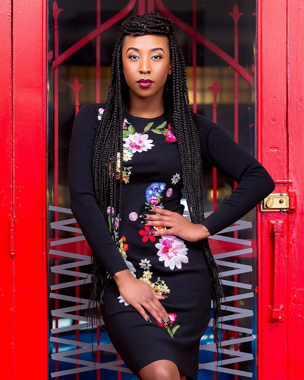 A woman with braided hair standing in front of a red door with a black and red metal security gate. She is wearing a black dress with colorful floral embroidery.