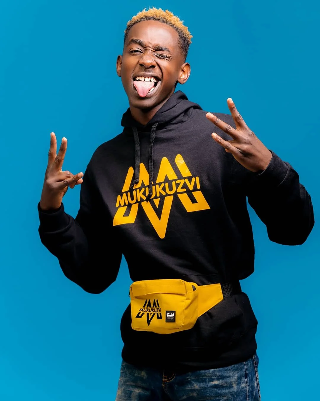 Young man with blonde hair making peace signs with both hands, sticking out his tongue, wearing a black hoodie with yellow letters and a yellow waist bag, standing against a blue background.