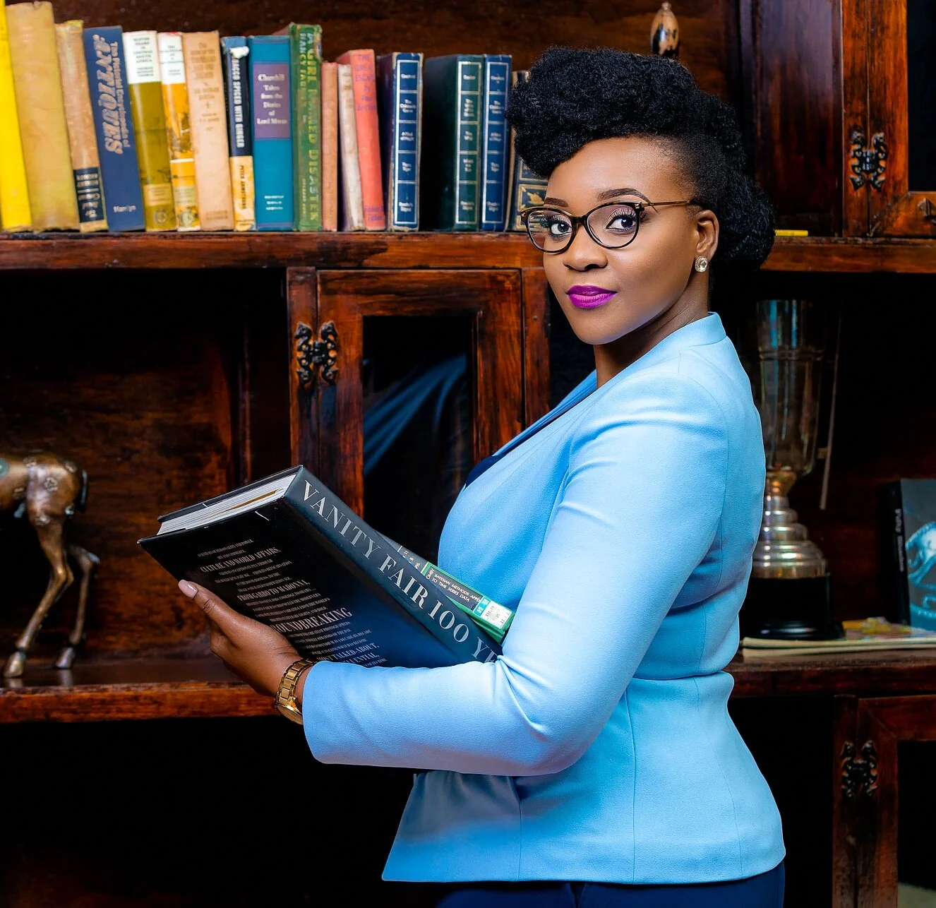 A professional woman with glasses and purple lipstick holding a book titled 'Vanity Fair 100 Years' in front of a wooden bookshelf filled with colorful books and decorative items.
