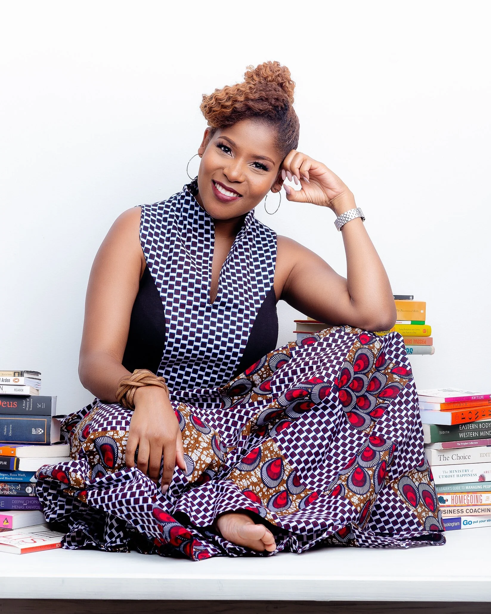 A smiling woman sitting on a white surface surrounded by stacks of books, wearing a patterned dress and accessories, with one hand resting on her face and the other on her lap.