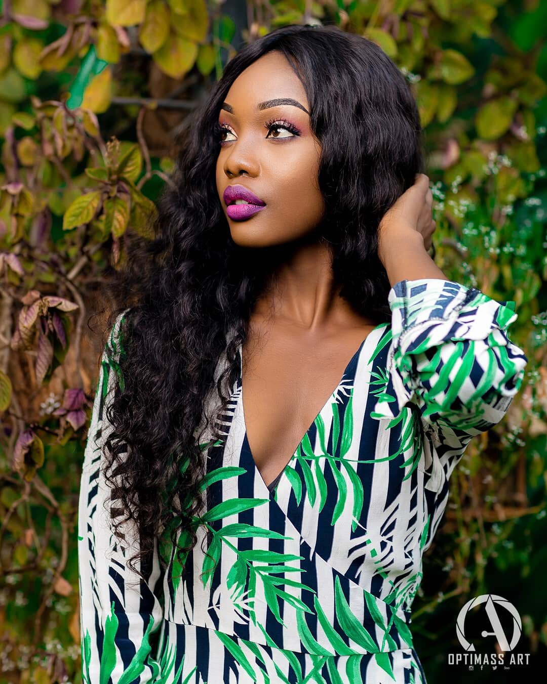 A woman with long curly black hair, wearing a green, white, and navy blue patterned dress, stands outdoors near green leafy plants, looking to her right.