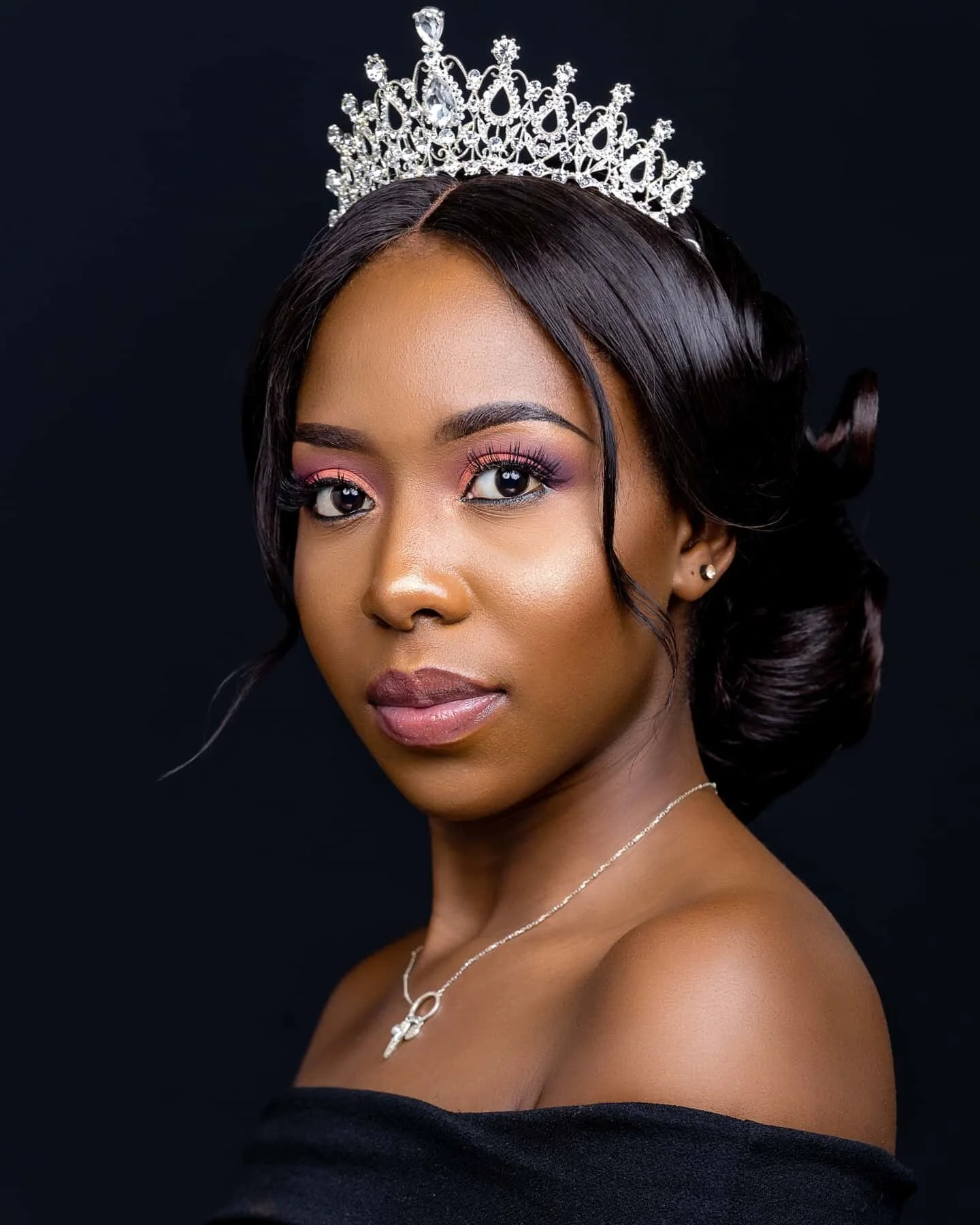 Portrait of a woman wearing a tiara, with styled black hair, pink eyeshadow, and a silver necklace, against a black background.
