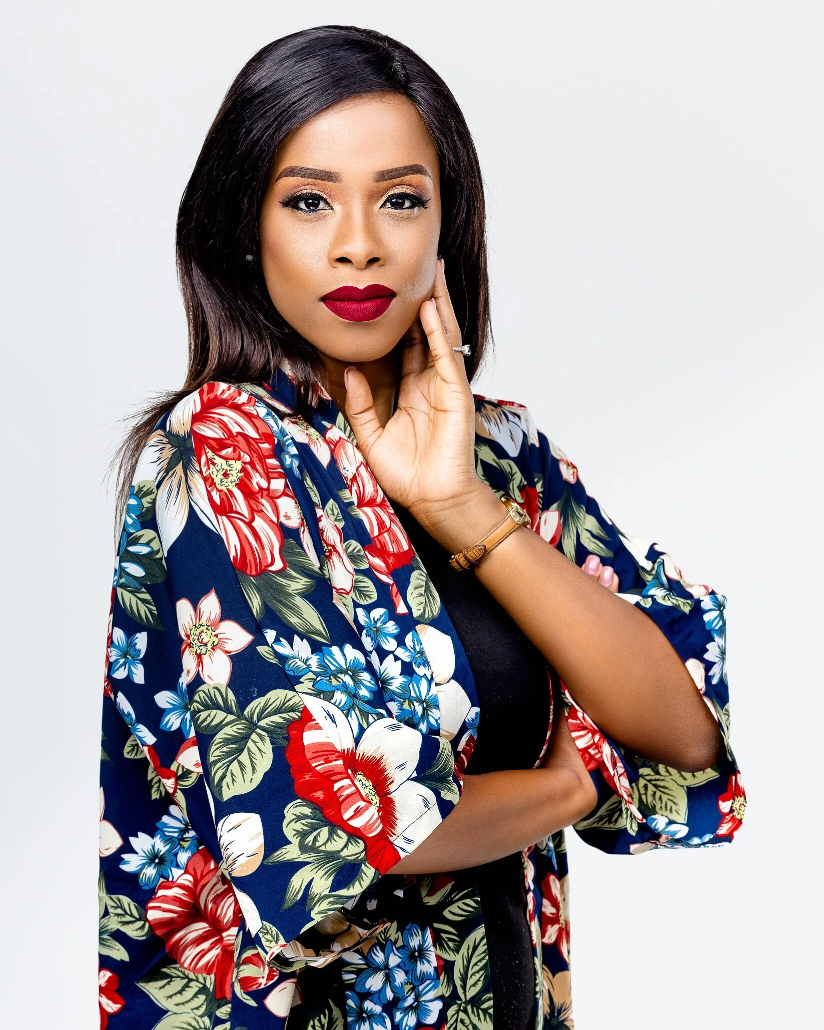 A woman with dark hair and red lipstick wearing a floral-patterned jacket over a black top, posing with her hand near her face against a plain white background.