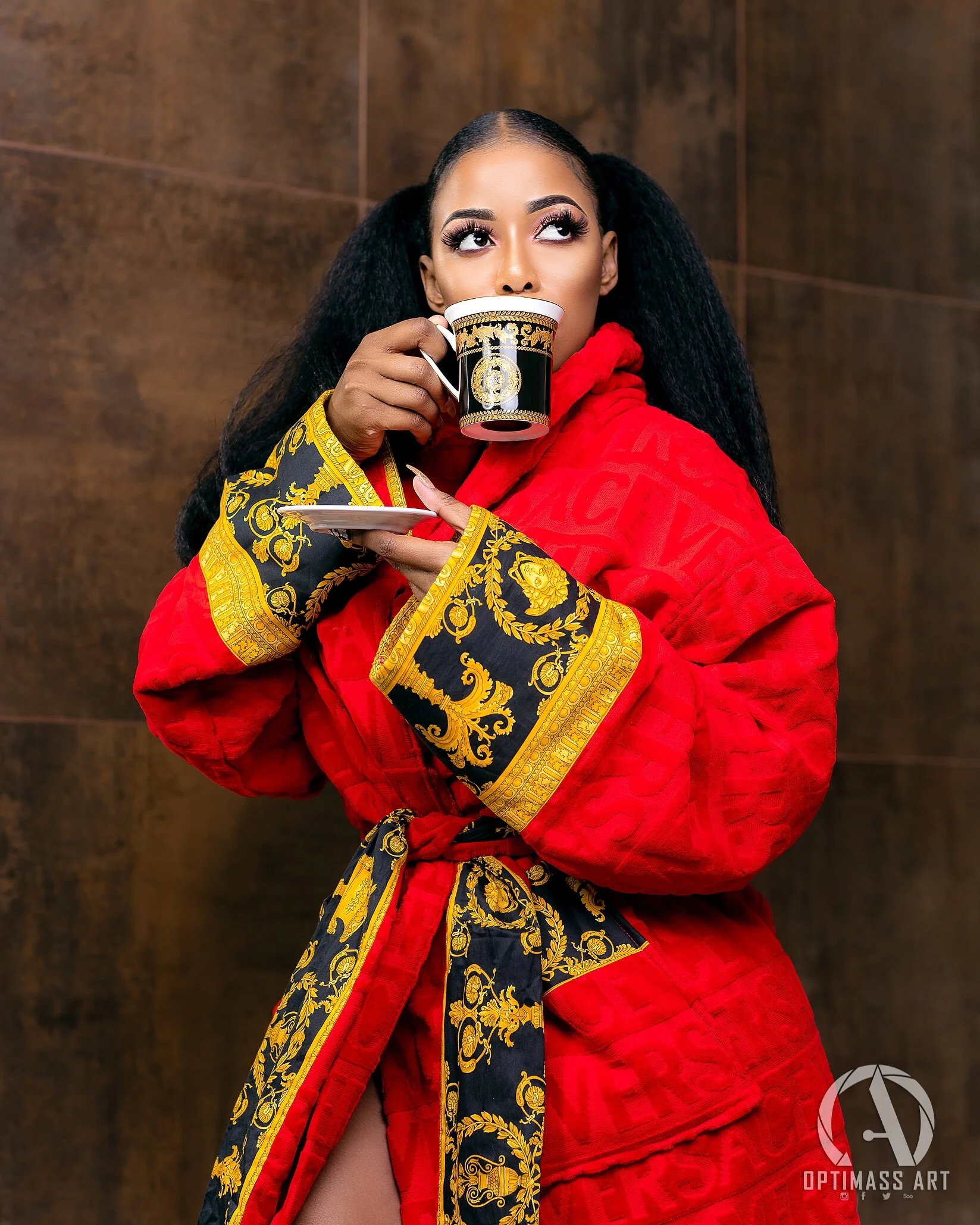 A woman with long dark hair styled in puffs is wearing a red quilted robe with black and gold ornate accents, and holding a black and gold cup with a saucer, standing against a brown background.