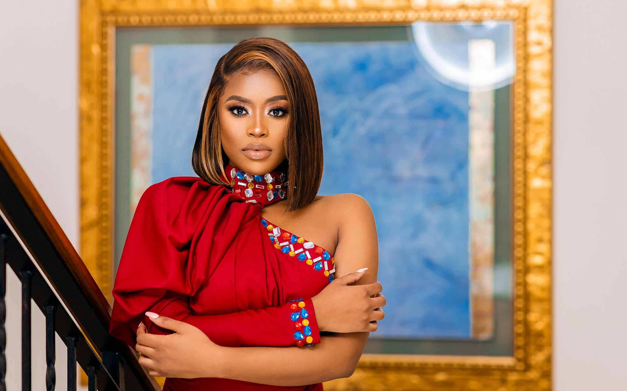 A woman with shoulder-length brown hair wearing a red dress with jeweled embellishments, posing indoors in front of a framed mirror.