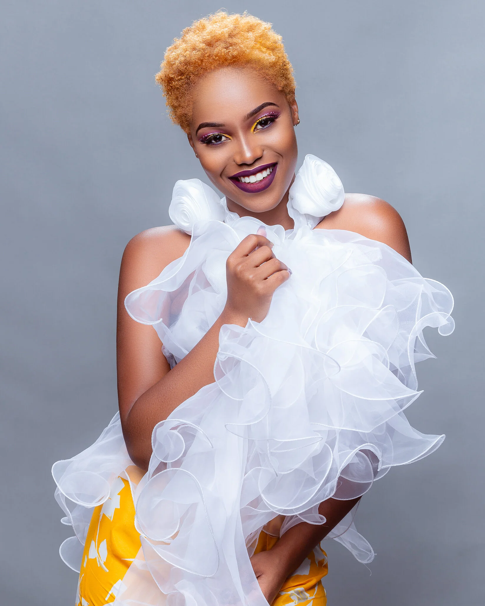 A woman with blonde curly hair, wearing bold makeup, smiling, holding a white ruffled fabric, dressed in yellow patterned pants, against a gray background.