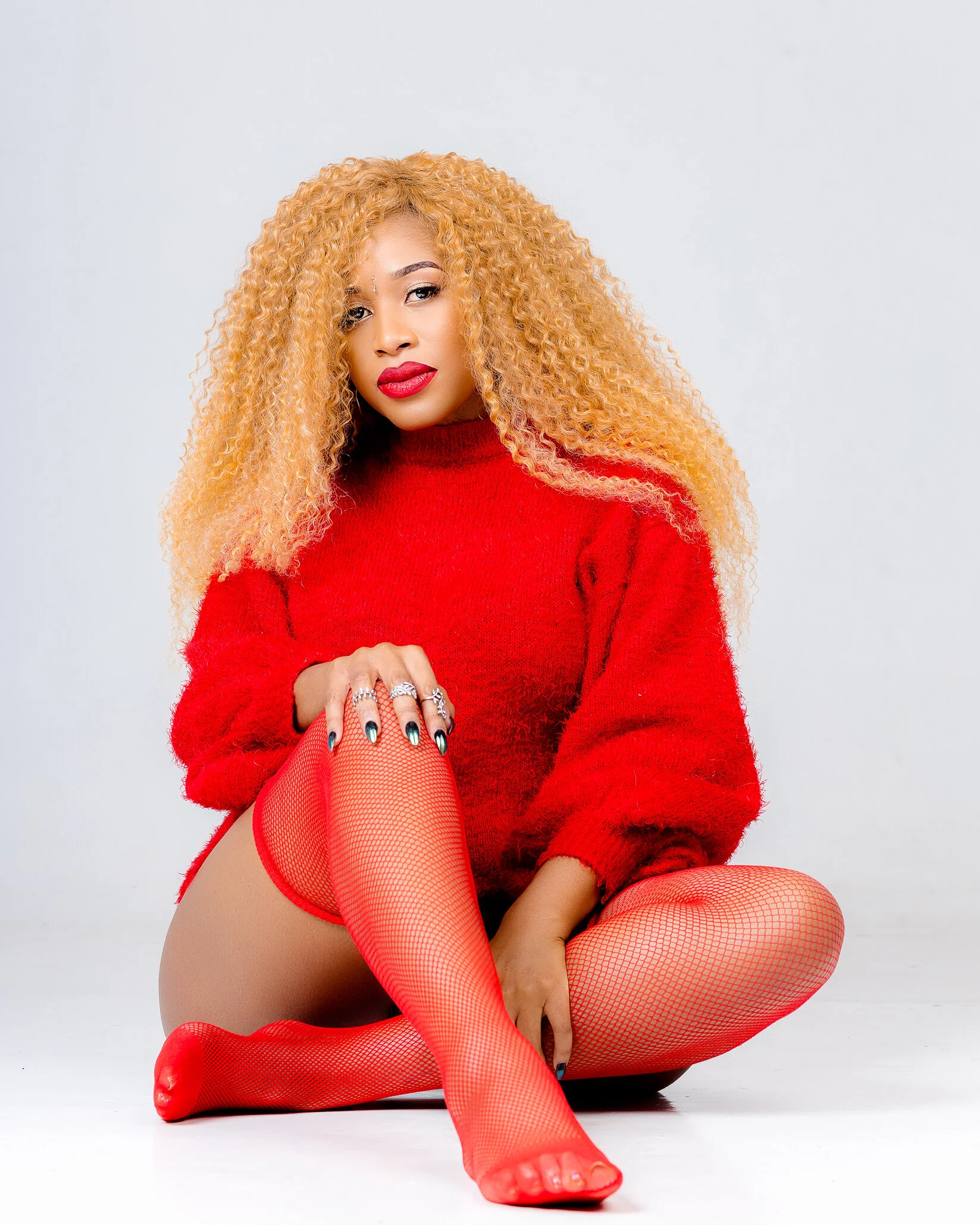 A woman with curly blond hair wearing a red sweater, red fishnet stockings, and red lipstick poses sitting on the floor against a plain white background.