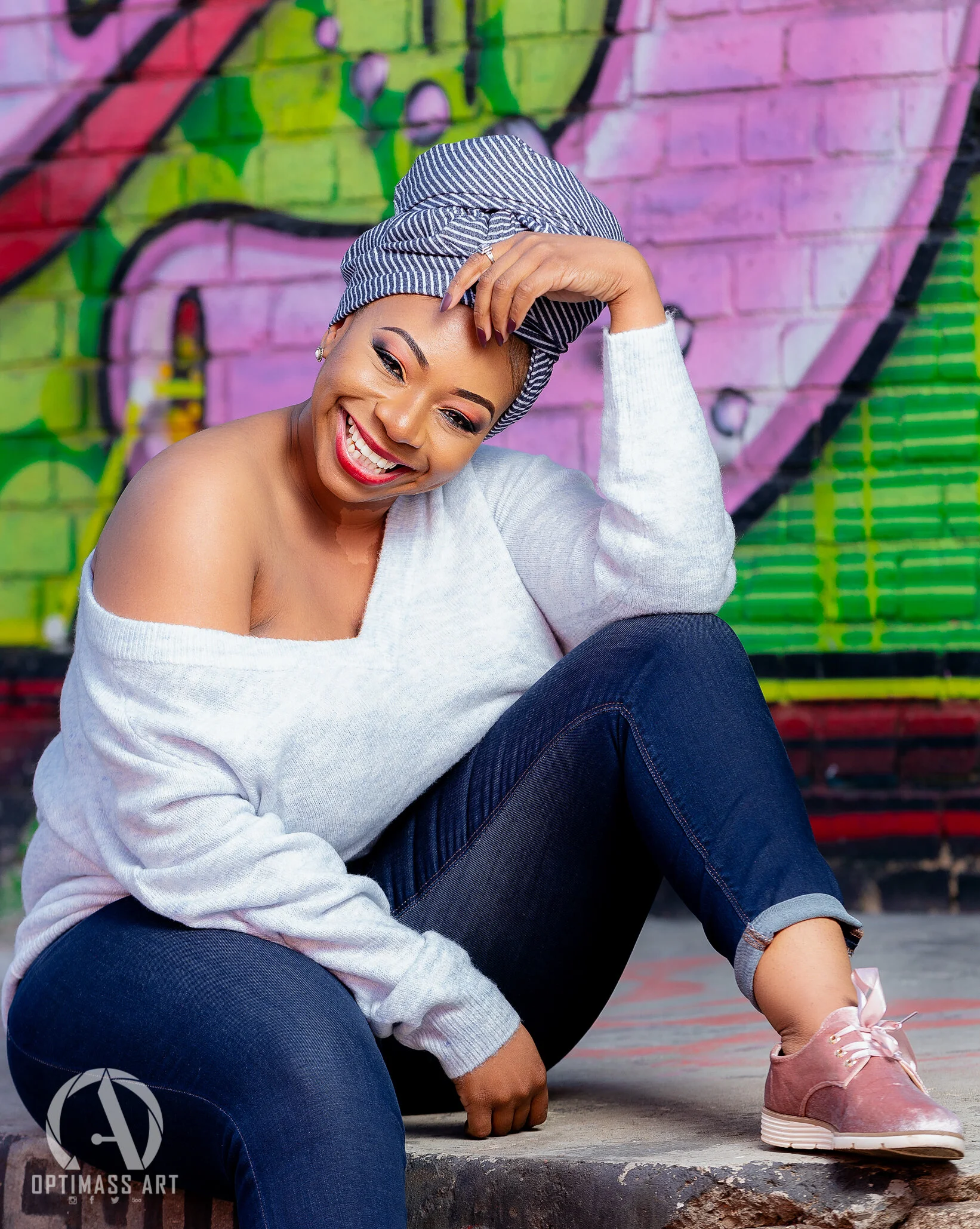 A woman sitting on the ground in front of a colorful graffiti wall, smiling and wearing a striped headscarf, white off-shoulder sweater, dark jeans, and pink sneakers.