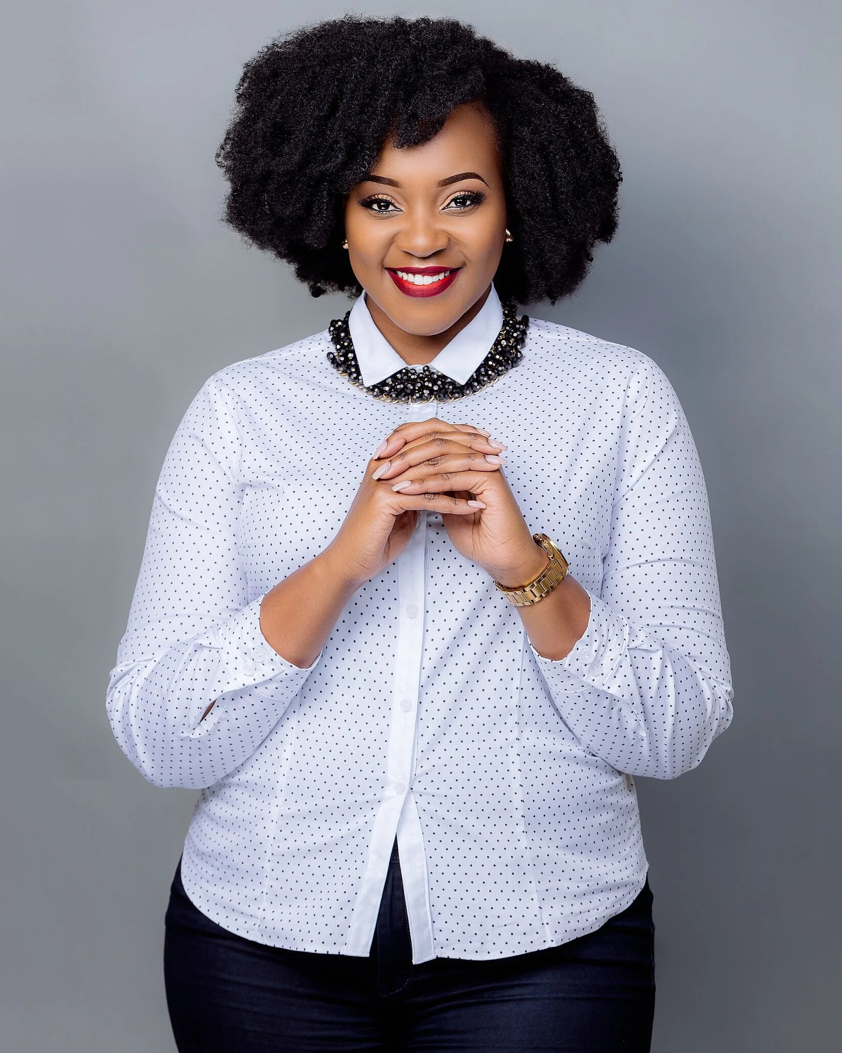 A woman with dark curly hair and red lipstick smiling, wearing a white polka dot blouse and a beaded necklace, posing with her hands clasped. Gray background.