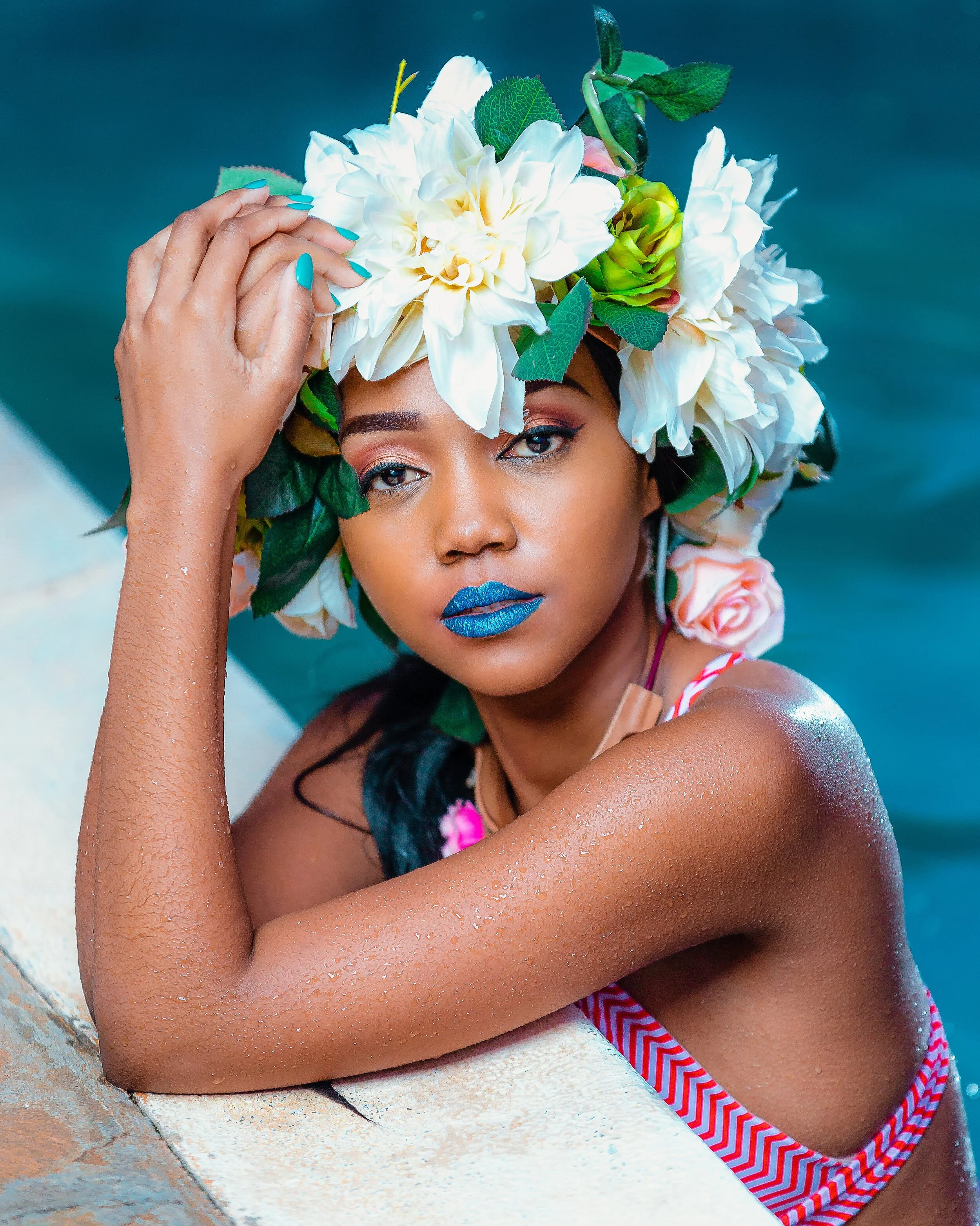 Woman with tan skin wearing a floral headpiece, blue lipstick, and a striped swimsuit, leaning on poolside edge.