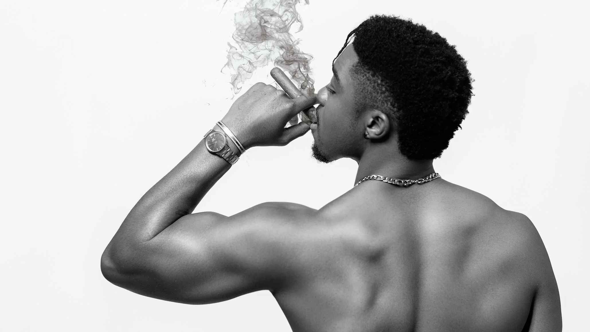 A shirtless young man with a side profile, short curly hair, a chain necklace, and a watch on his wrist, smoking a cigar, with smoke rising from it, against a plain background.