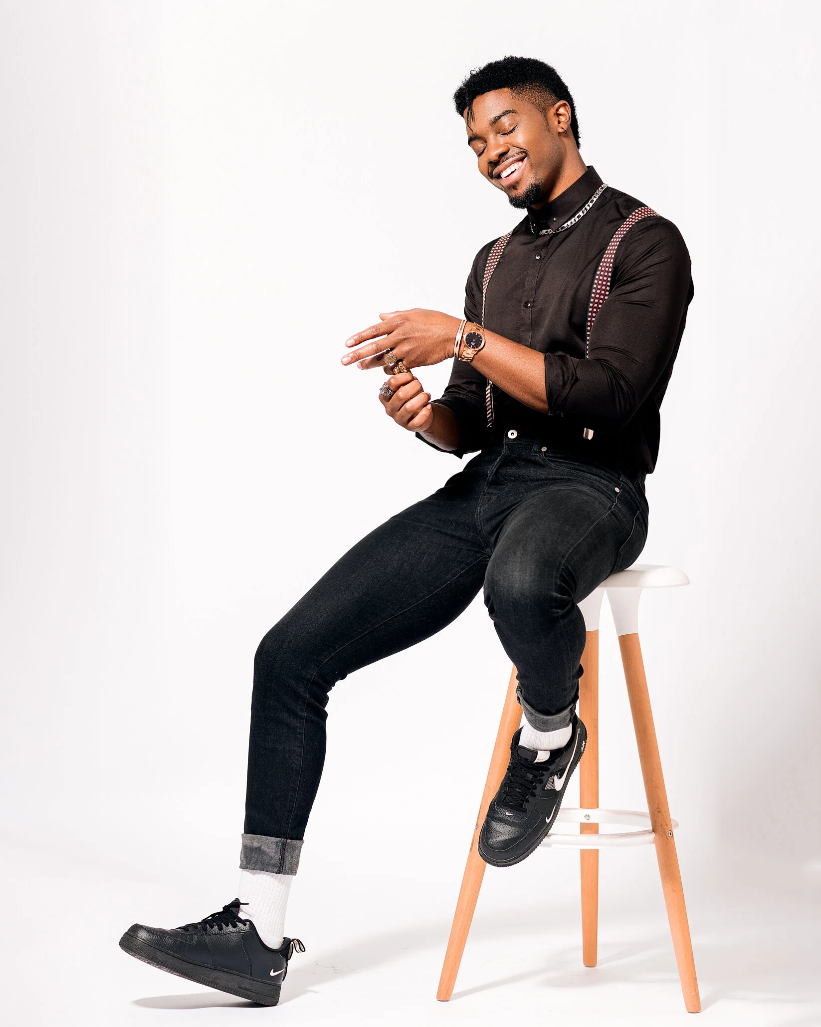 A smiling man with dark hair and dark skin sitting on a white stool against a white background, wearing black jeans, a black shirt, black sneakers, and accessories.