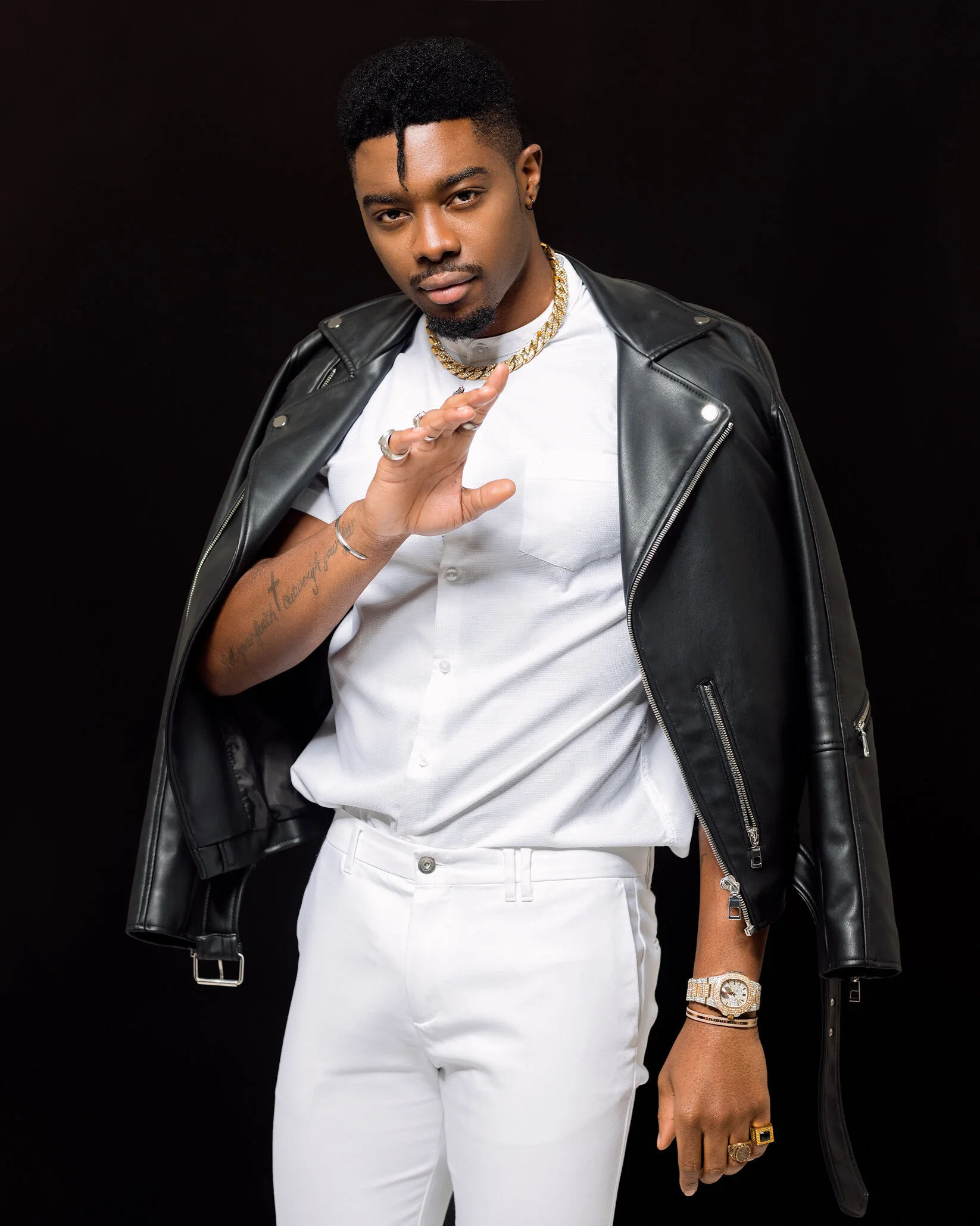 A young man with dark skin, wearing a black leather jacket draped over his shoulders, a white button-up shirt, white pants, and gold jewelry, posing against a black background.