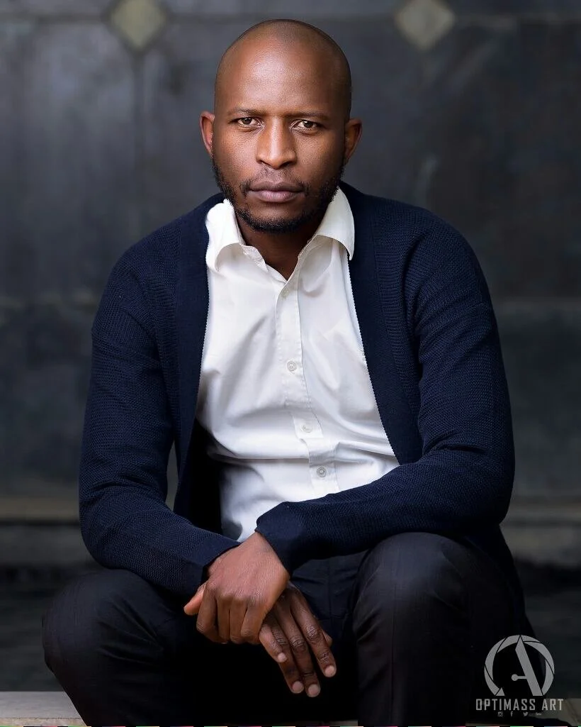 A man with a shaved head and beard sitting with his hands clasped, wearing a white collared shirt and navy blue cardigan, looking directly at the camera against a dark, blurred background.