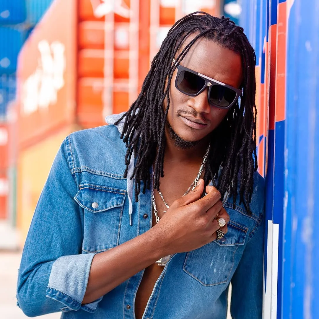 A young man with dreadlocks wearing sunglasses, a denim jacket, and jewelry, leaning against colorful lockers.