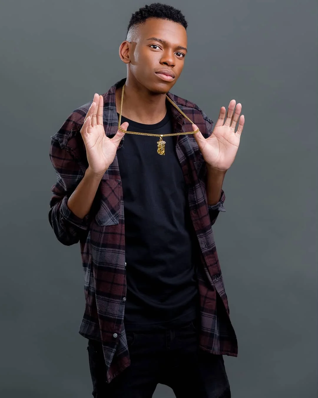 A young man with short curly hair wearing a black T-shirt, an open plaid shirt, and holding up a gold chain with a pendant.