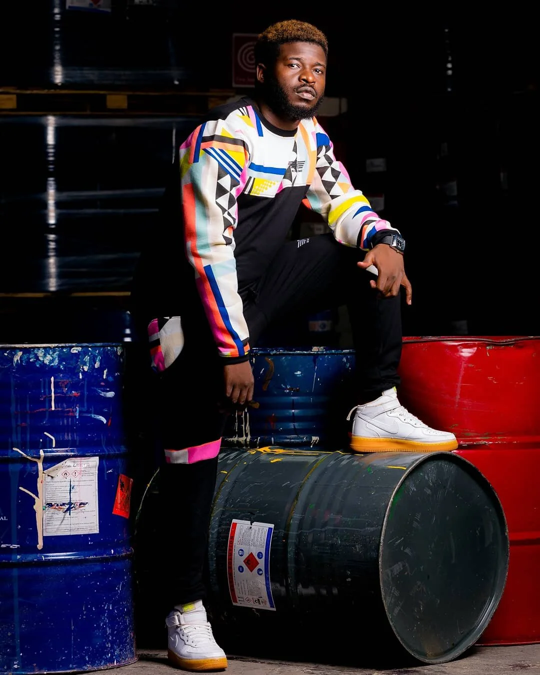 A man with a beard and styled hair, wearing a colorful geometric-patterned jacket, black pants, and white sneakers, sits on a stack of industrial barrels in a dark setting.