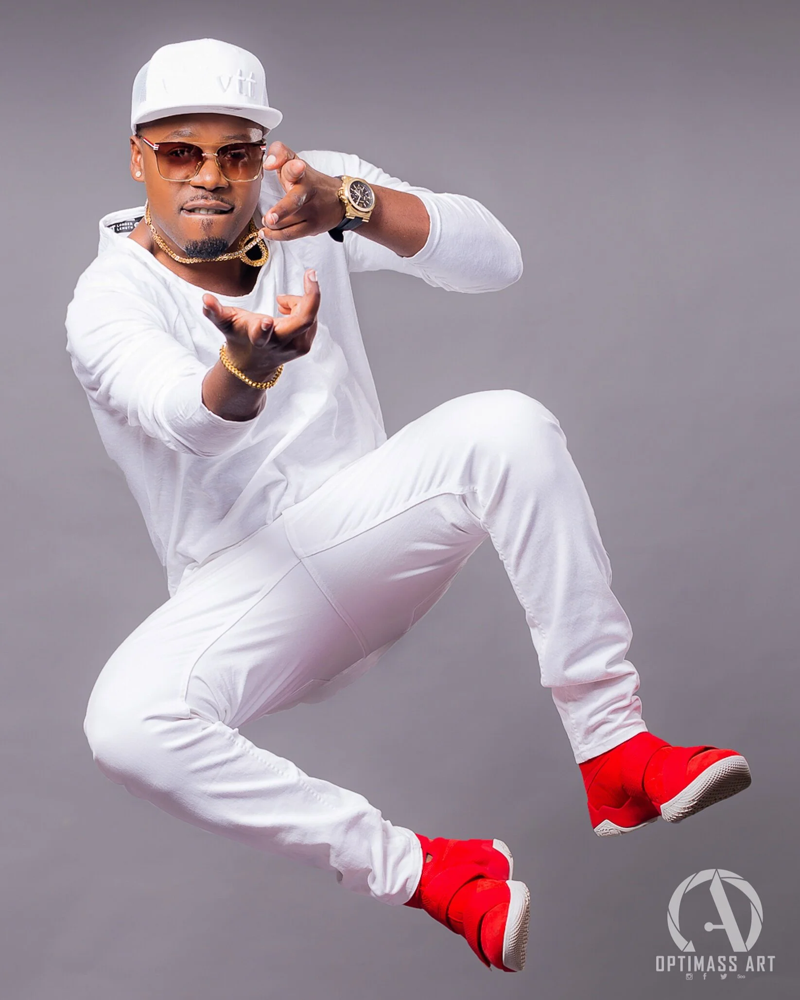 Man wearing white clothing, red sneakers, sunglasses, and accessories, jumping in front of a gray background.