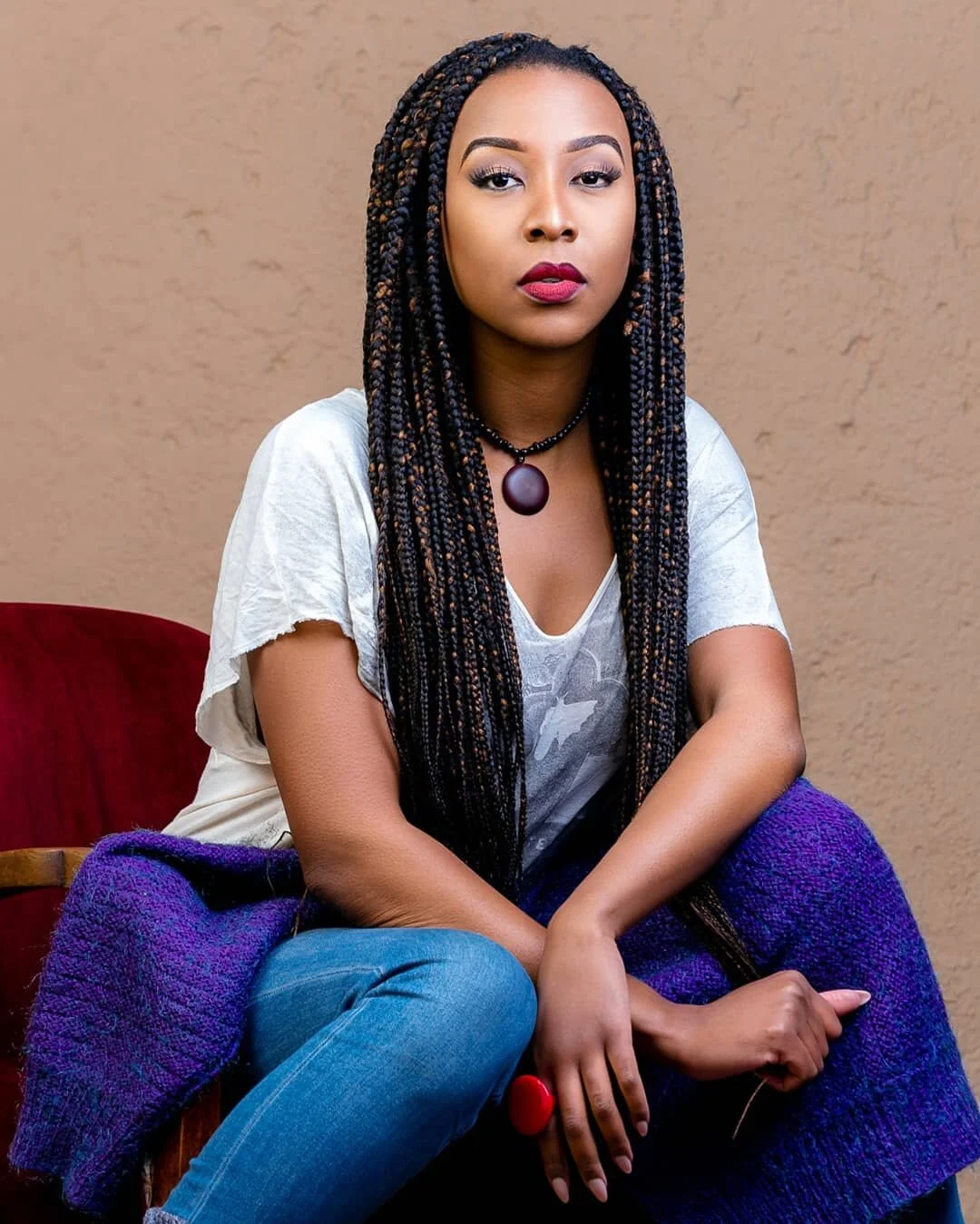 A woman with long braided hair, wearing a white t-shirt, a black beaded necklace with a large pendant, and blue jeans, sitting on a chair with a purple blanket draped over her lap, against a beige wall.