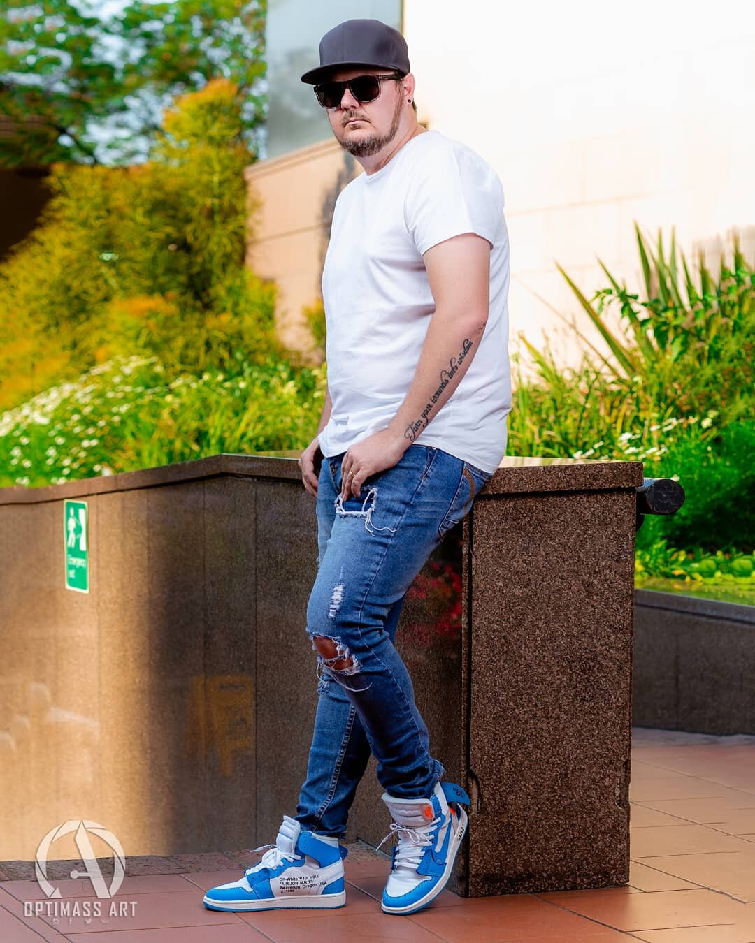 A man with sunglasses, a black baseball cap, a white T-shirt, and ripped blue jeans leaning against a brown concrete ledge outdoors, with green foliage in the background.