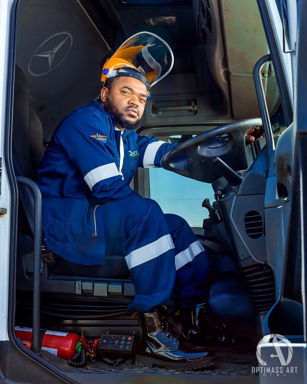 A man in a blue work uniform sitting in the driver's seat of a truck, wearing a yellow safety helmet with a face shield. The interior of the truck shows a Mercedes logo on the wall and an emergency fire extinguisher and a radio on the floor.