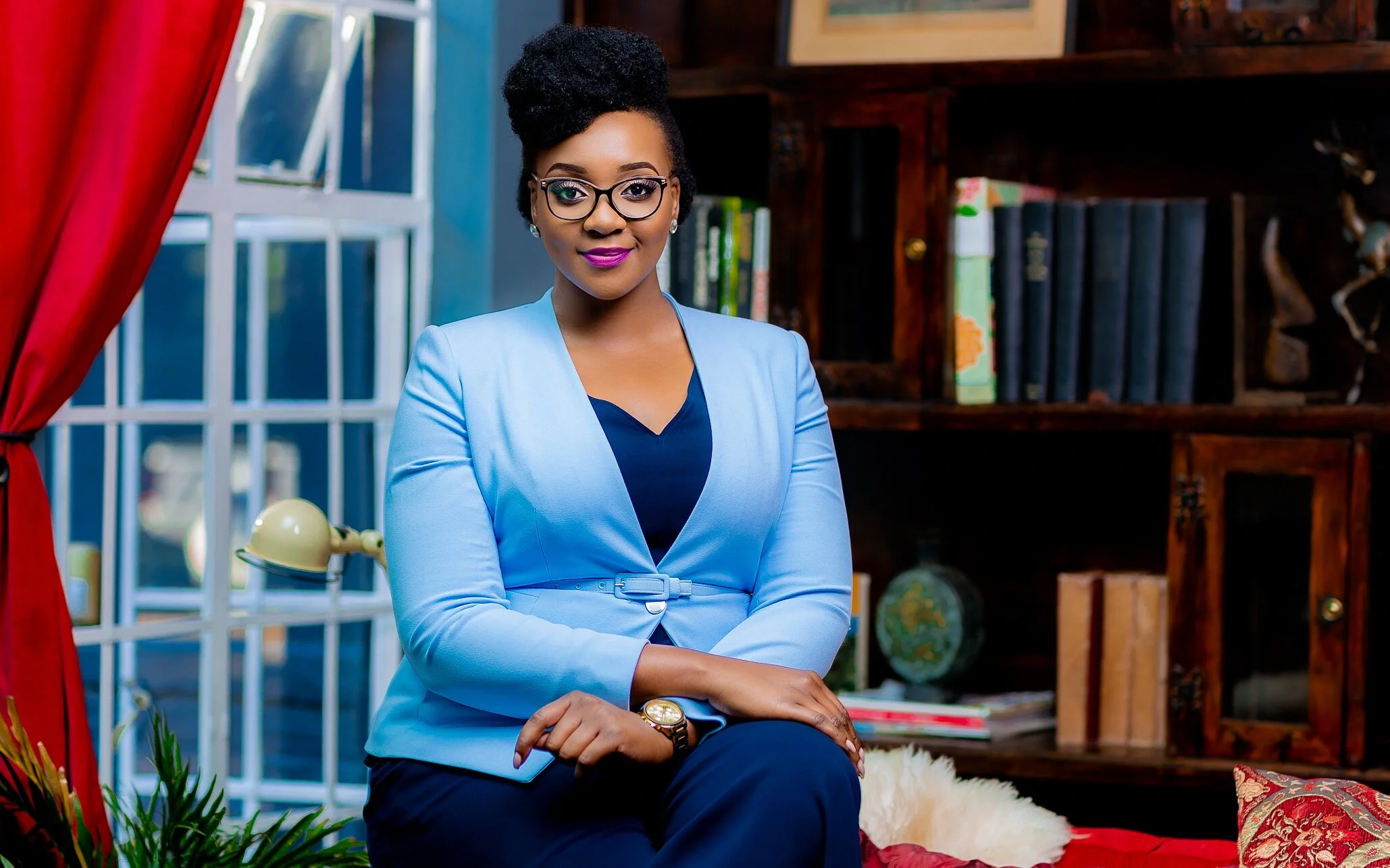 A professional woman with glasses and purple lipstick sitting confidently in a well-decorated room with bookshelves, a window with red curtains, a lamp, and a sofa.