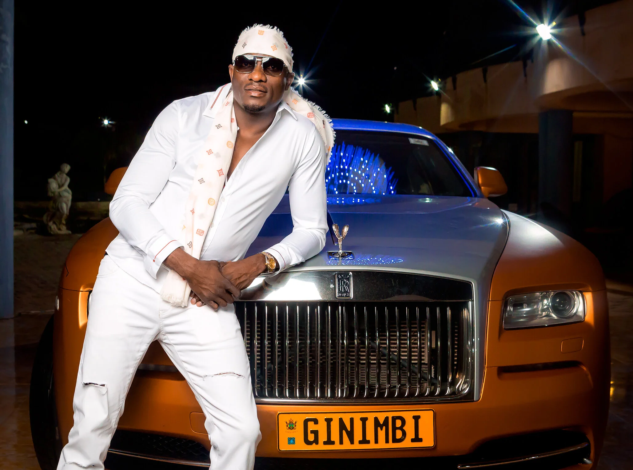 A man in a white outfit and sunglasses leaning against an orange Rolls Royce with a customized license plate reading 'GINIMBI' at night, with a dark background and decorative lighting.