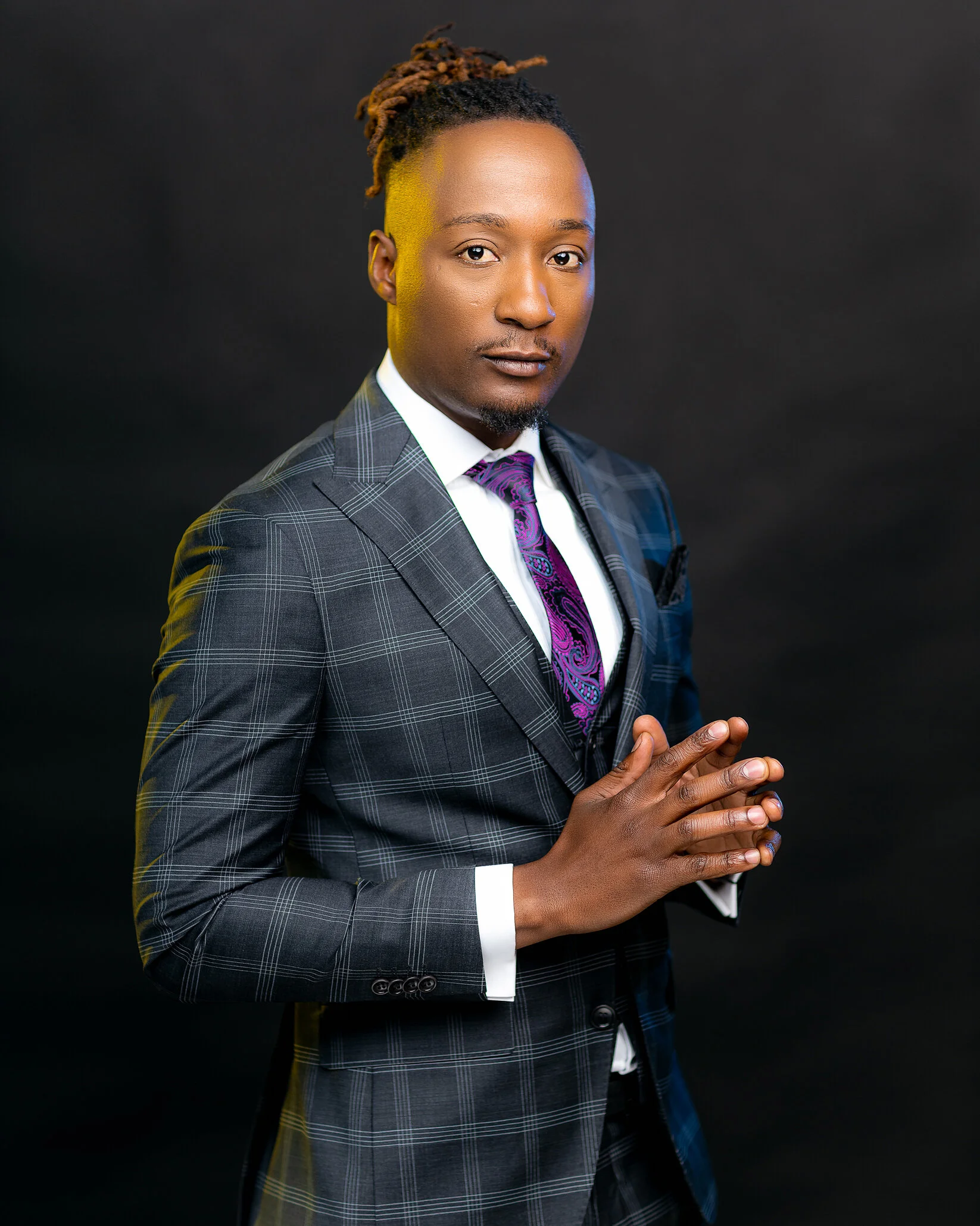 A young man wearing a gray plaid suit with a white shirt and a purple paisley tie, posing with hands pressed together in front of a black background.