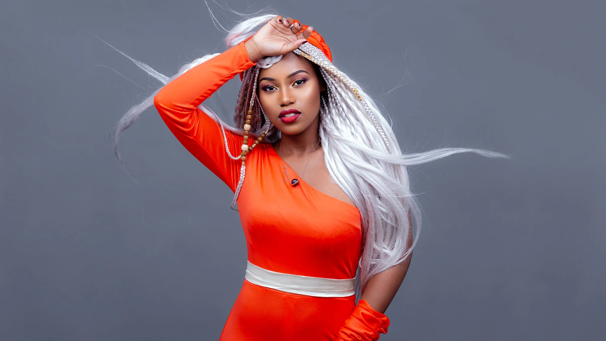 A woman with long white braided hair wearing an orange dress with a white waistband, posing with her hand on her head against a gray background.