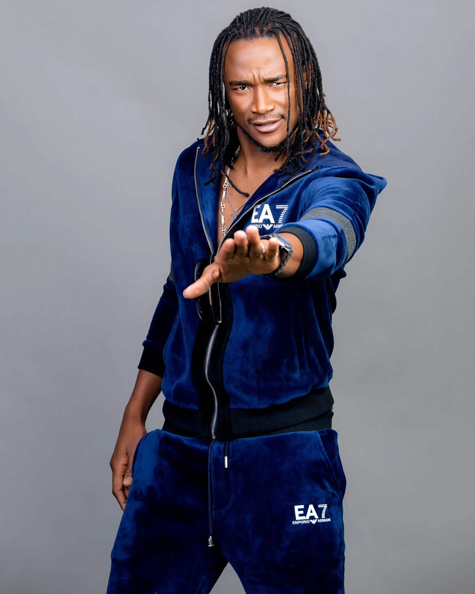 Man with dreadlocks wearing a blue velvet tracksuit with white EA7 and Emporio Armani logos, posing with one hand extended forward