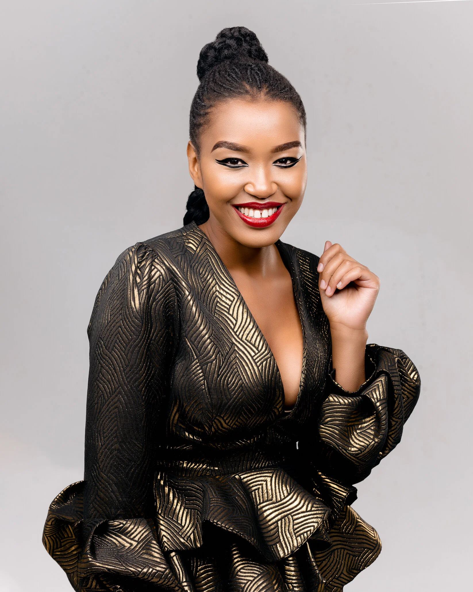 A woman with braided hair styled in a high bun, wearing a black and gold patterned dress with puffed sleeves, smiling with bright red lipstick, against a plain gray background.