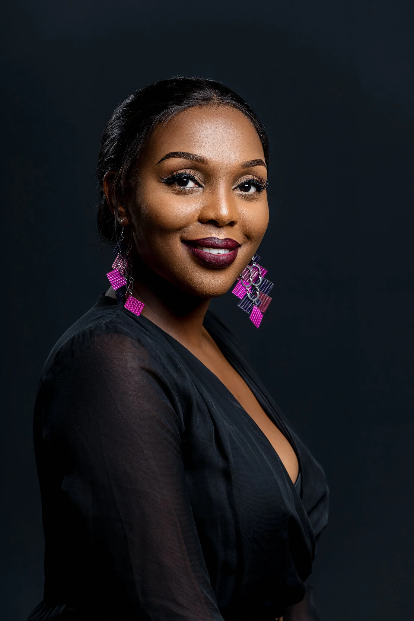 Portrait of a woman with dark hair styled back, wearing bold earrings and makeup, dressed in black, against a dark background.