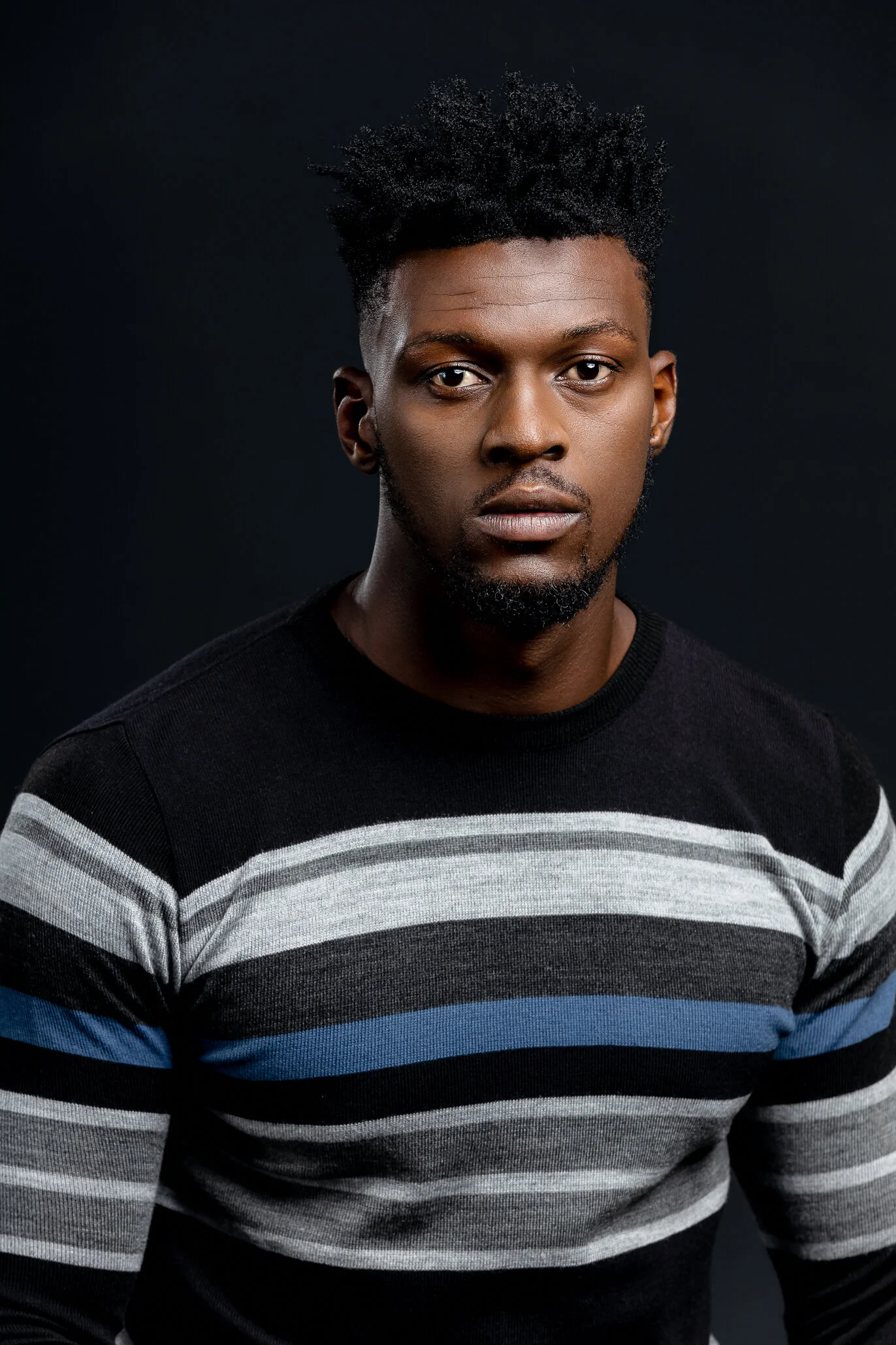A young man with short, curly hair and a beard, wearing a black, gray, white, and blue striped sweater, posing against a dark background.
