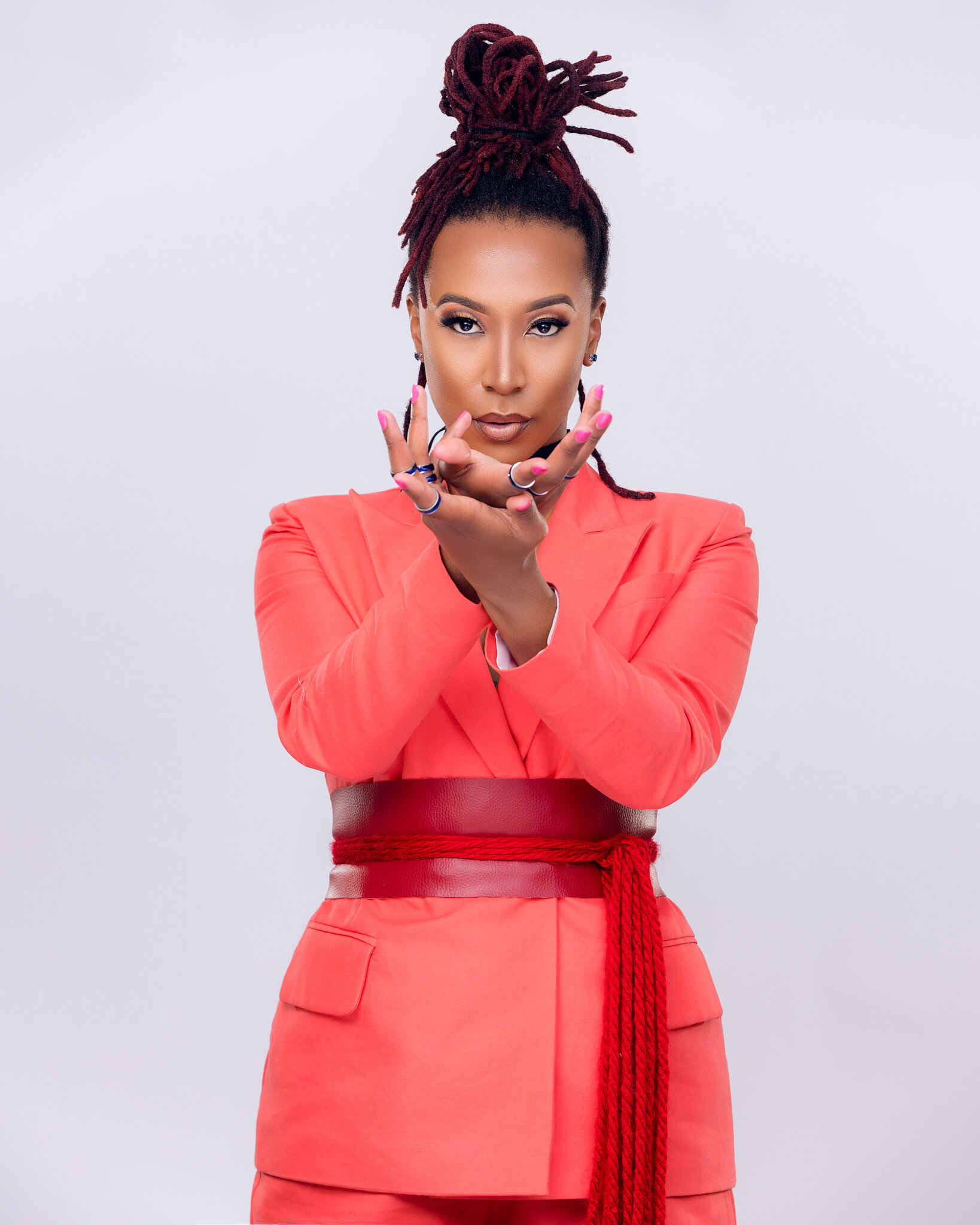 A woman with styled dreadlocks in a top knot, wearing a coral blazer and matching skirt with red belt and sash, making a gesture with her hands.