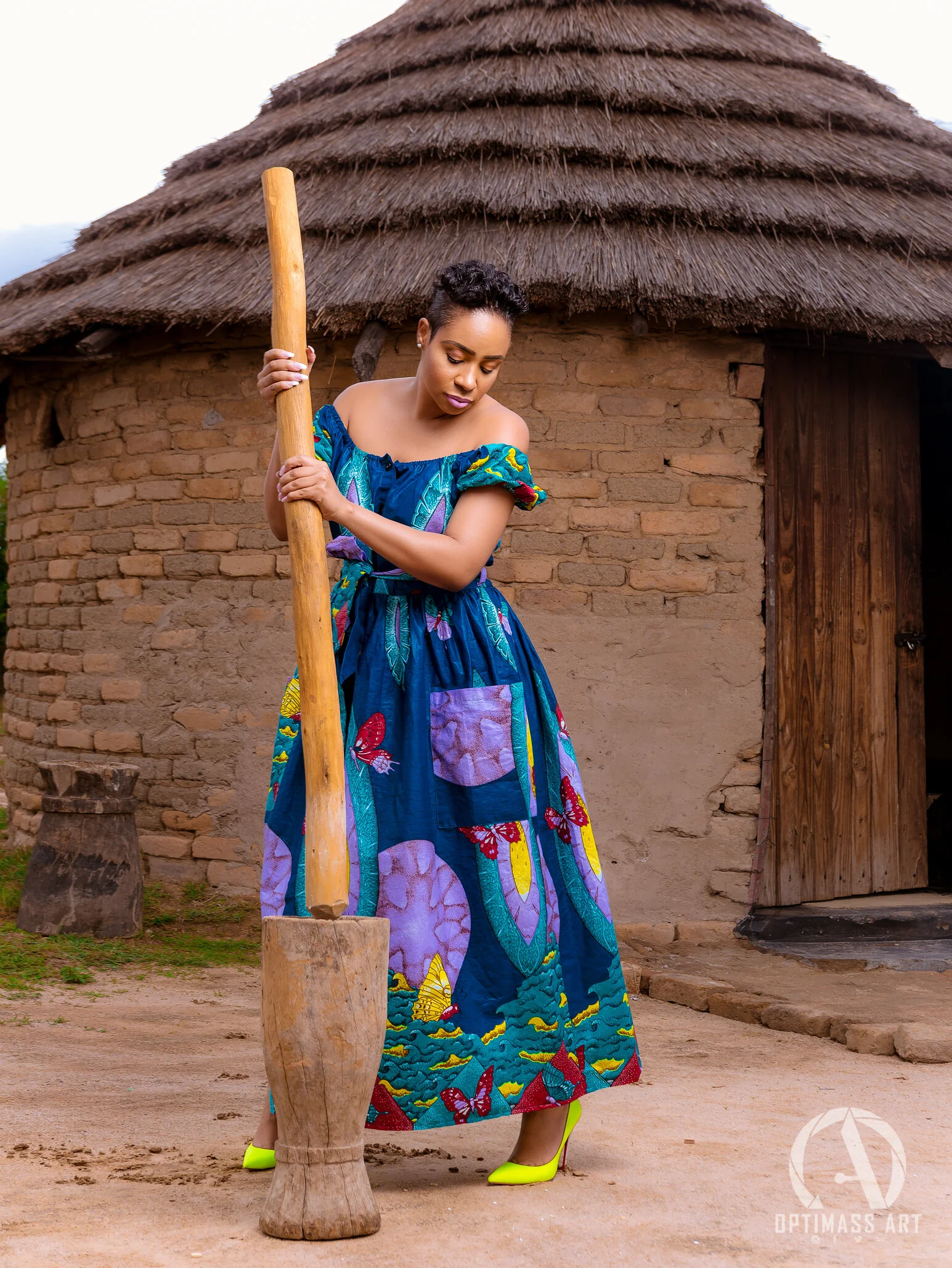 A woman in colorful African print dress standing outside a traditional mud and thatch hut, holding an ax.