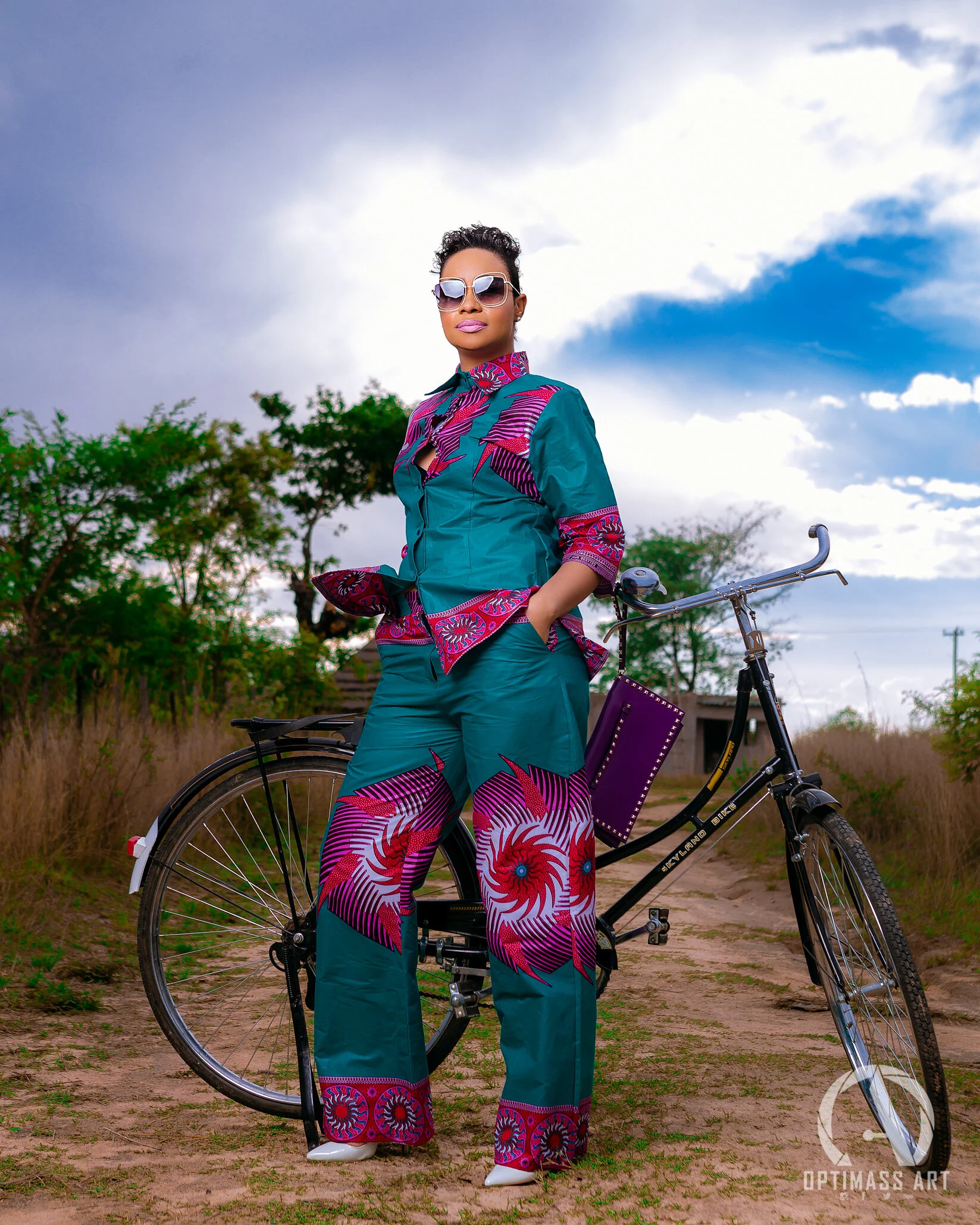 Woman standing outdoors with hands in pockets, wearing sunglasses, a teal and pink patterned outfit, and white shoes, beside a black bicycle on a dirt path with trees and cloudy sky in the background.