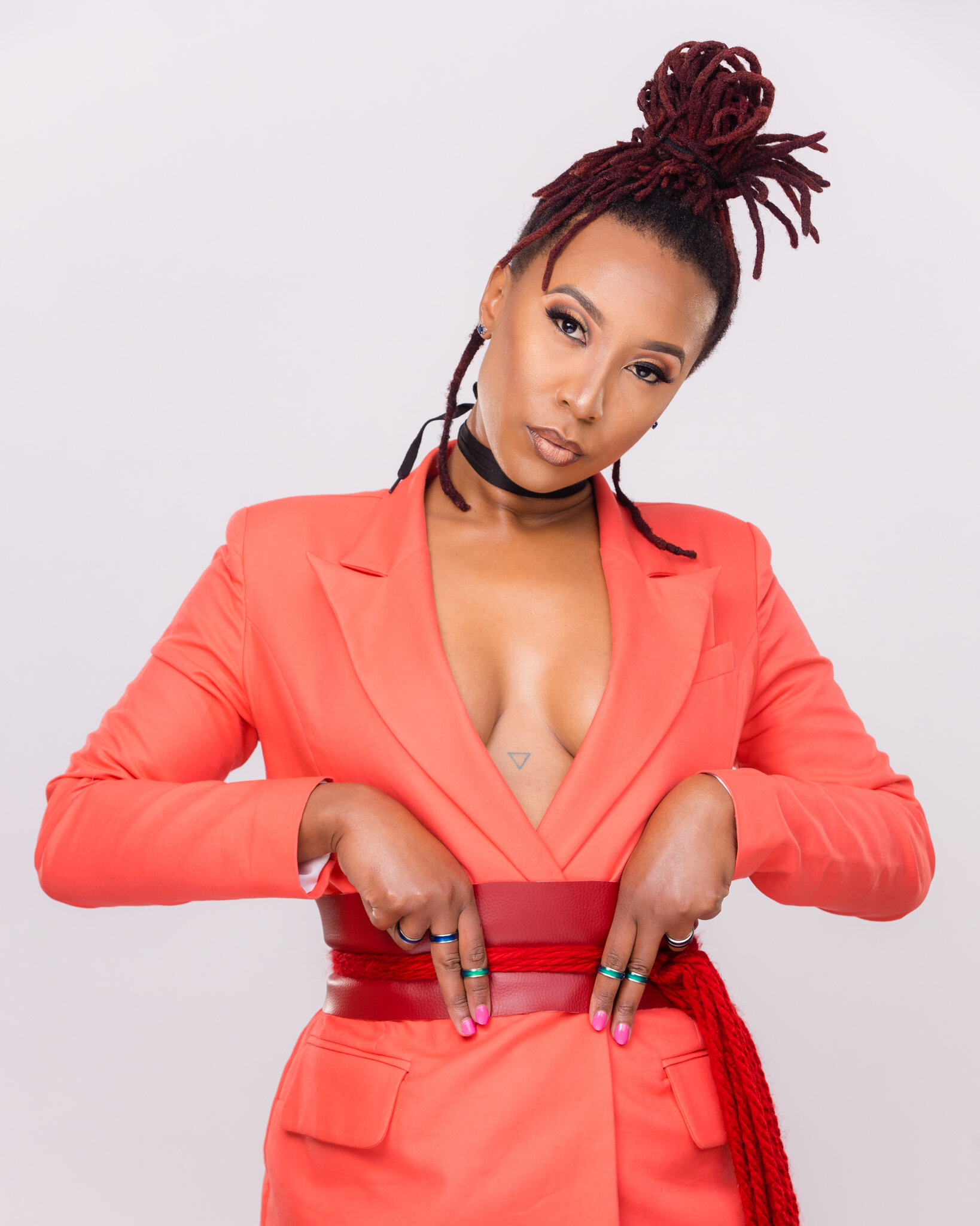 Woman with red dreadlocks tied in a high bun, wearing a coral blazer and black choker, standing against a plain white background.