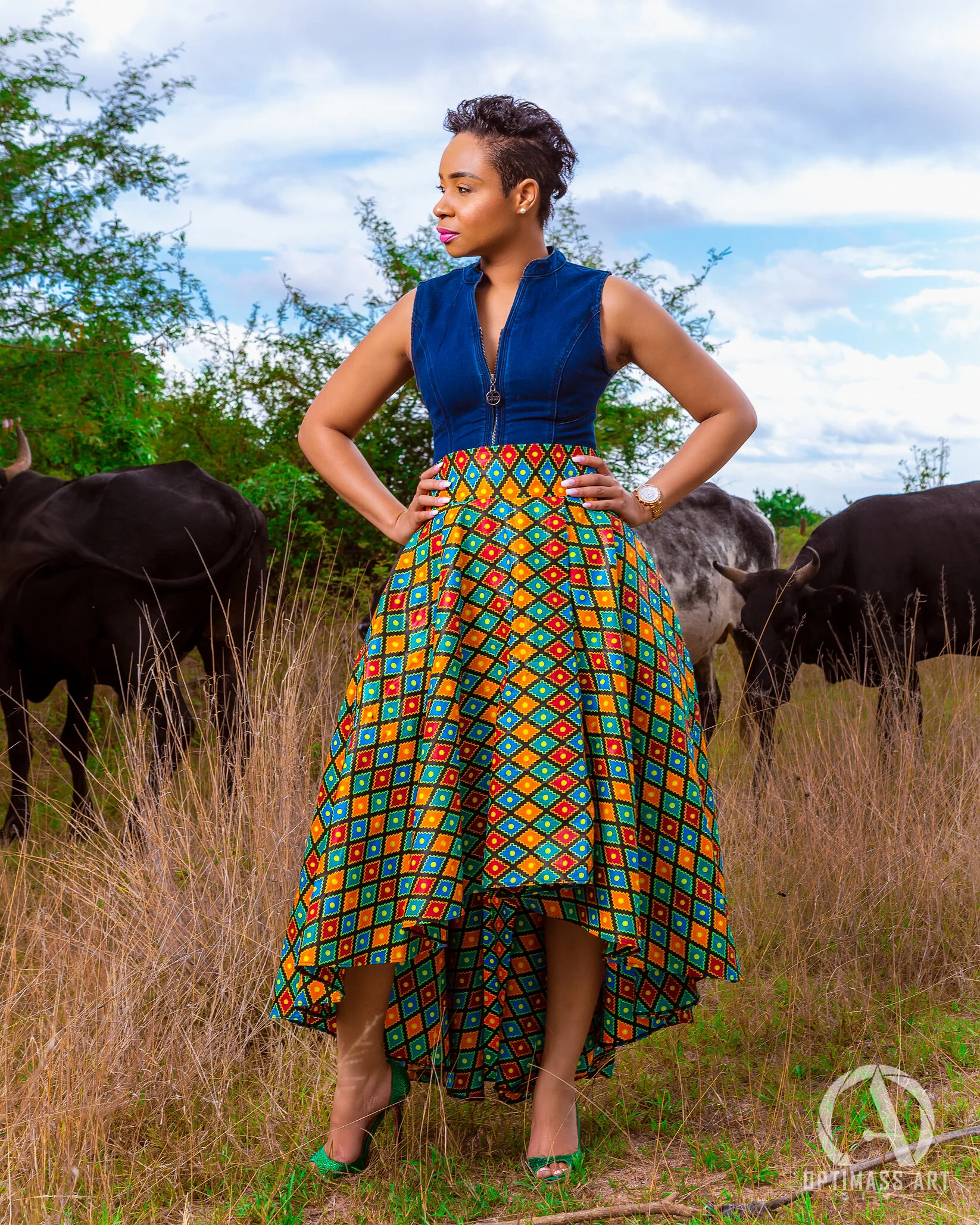 A woman in a blue sleeveless top and a vibrant, multicolored African print skirt standing in a grassy field with cows behind her, looking to the side.