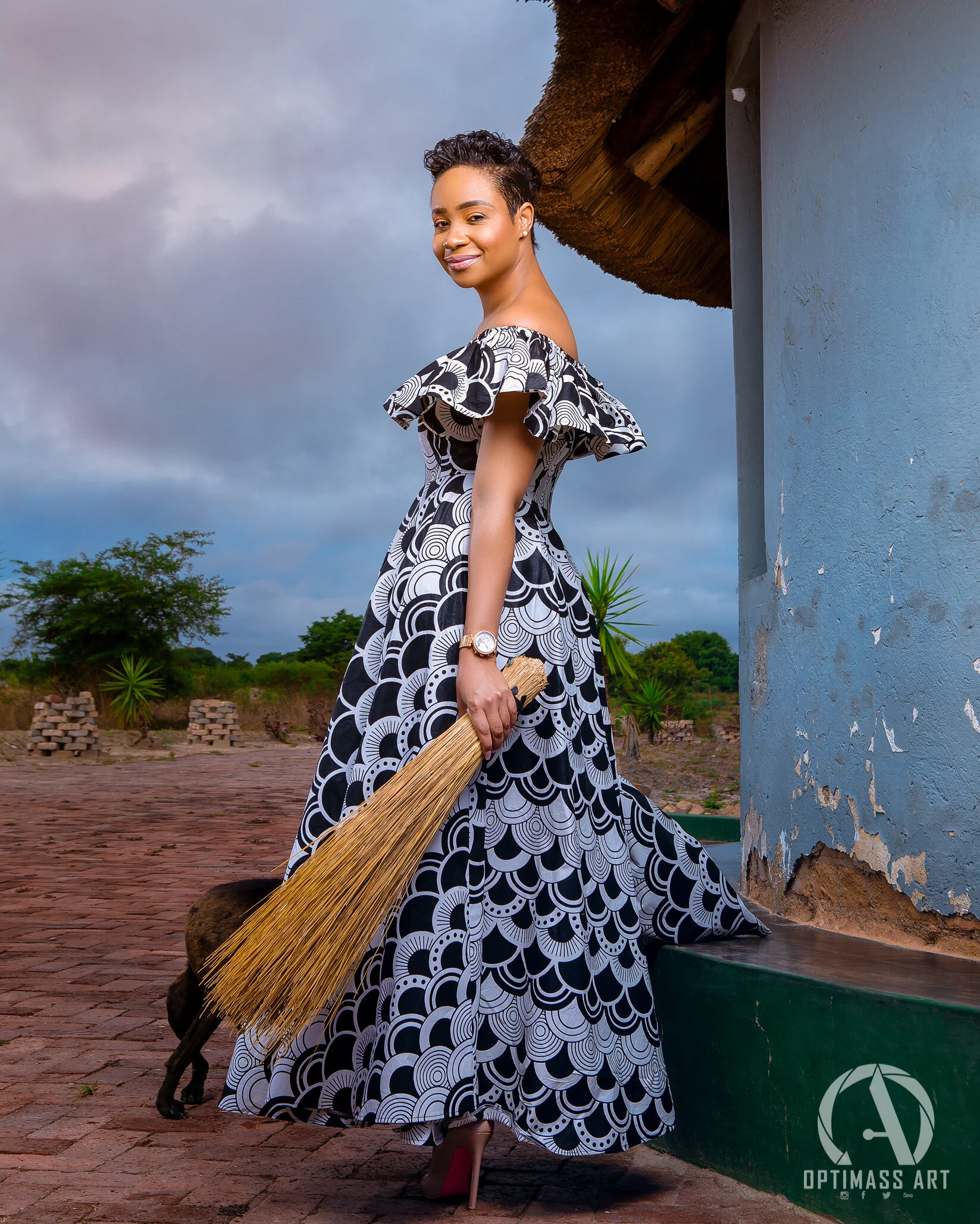 A woman wearing a black and white patterned dress, holding a broom, standing outdoors near a building with a thatched roof, with a cloudy sky and greenery in the background.