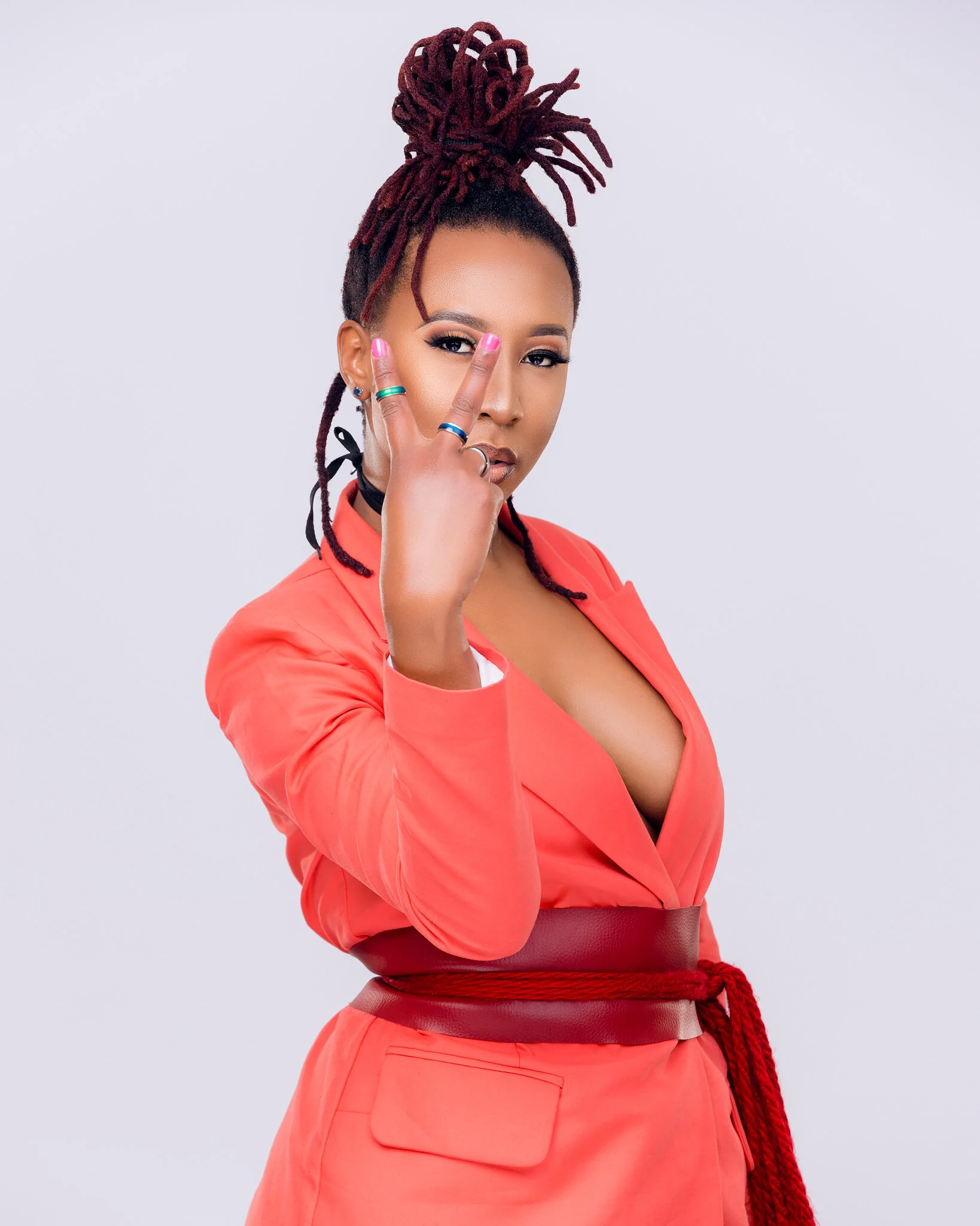 A woman with dreadlocks styled in a high bun wearing a coral-colored blazer and skirt, making a peace sign near her eye with her fingers, against a plain light background.