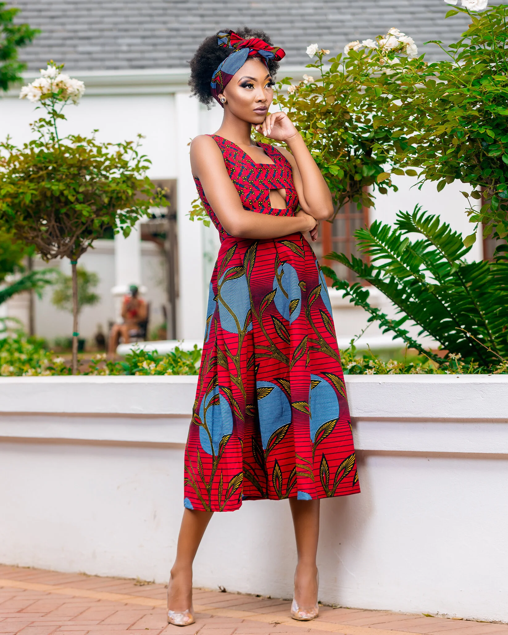 A woman in a colorful, patterned dress with a matching headband standing outdoors near green plants and flowers, with a person blurred in the background.