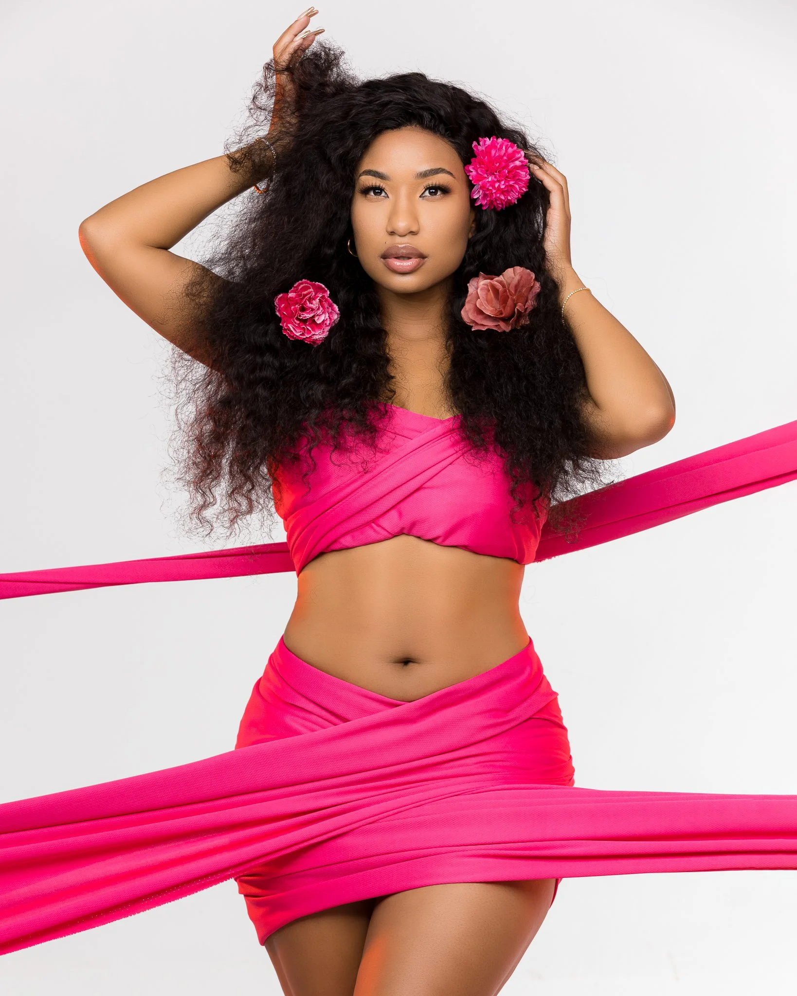 A young woman with long, curly black hair decorated with large pink flowers, wearing a bright pink strapless top and skirt, posing with pink fabric straps, against a plain white background.